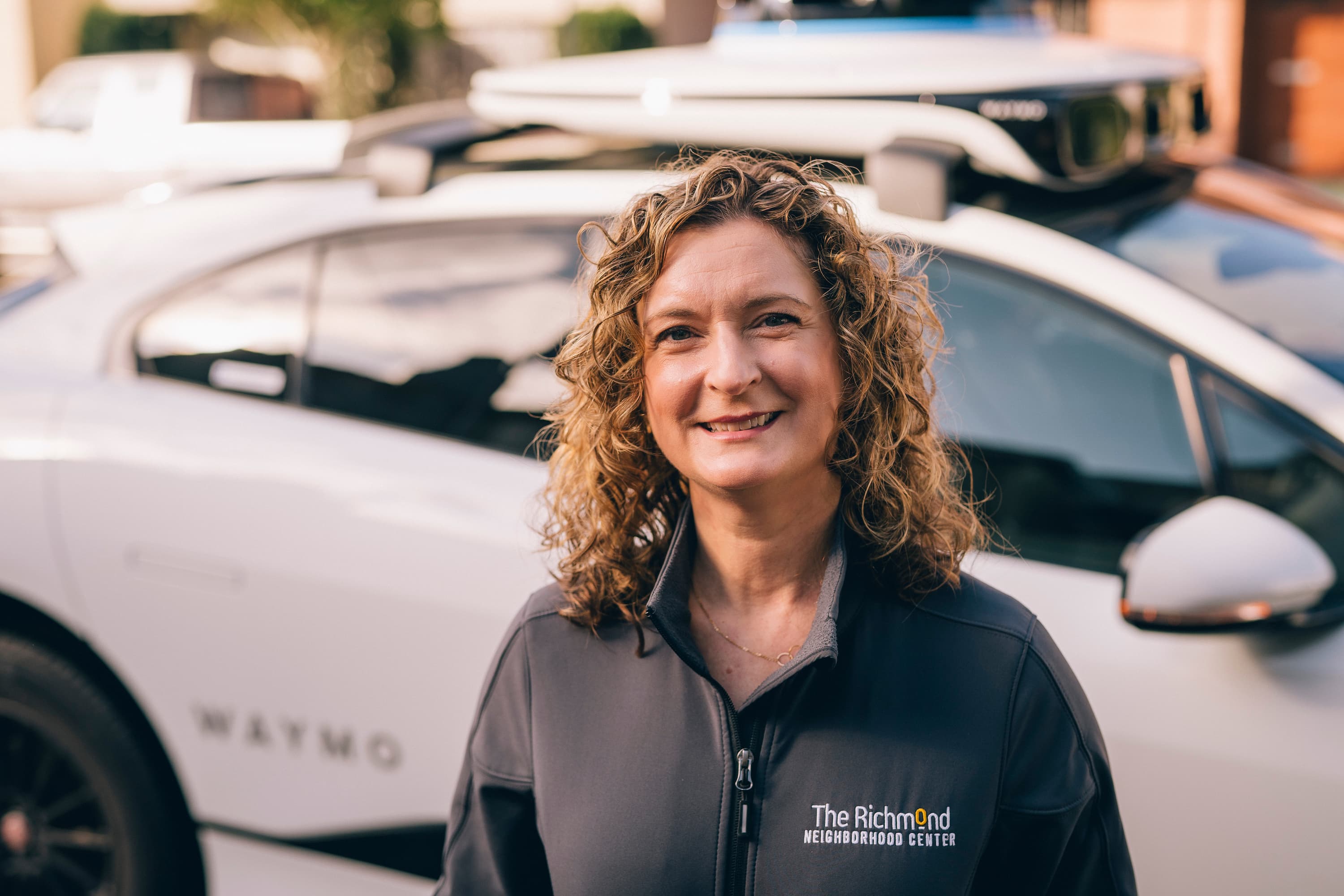 A smiling woman with curly hair, in front of a white car with the word &quot;WAYMO&quot; on it.