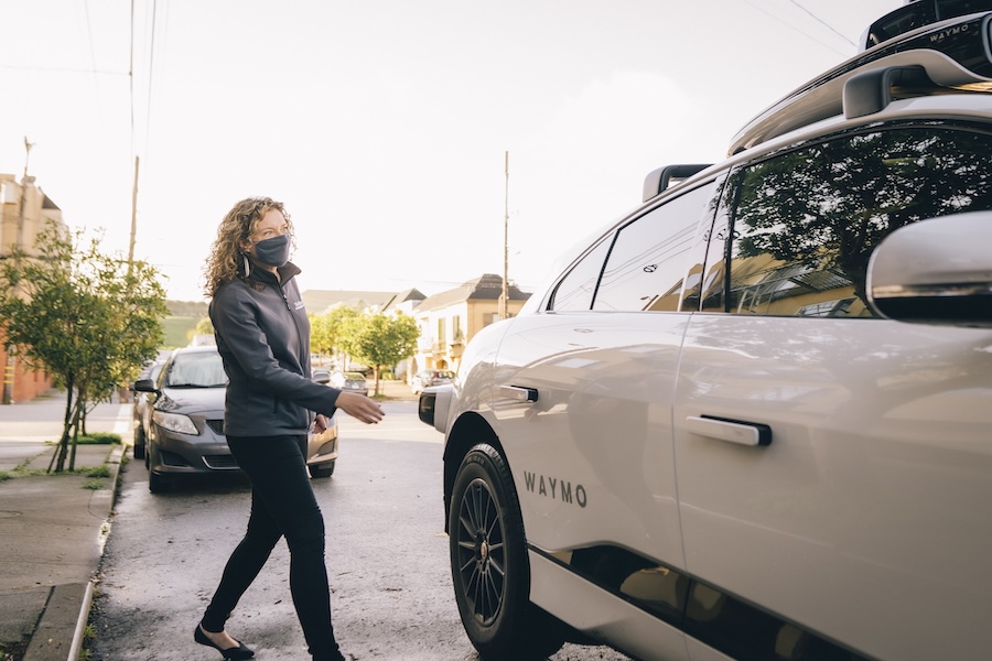 A person with a face mask approaches a white Waymo autonomous vehicle on a sunny street.