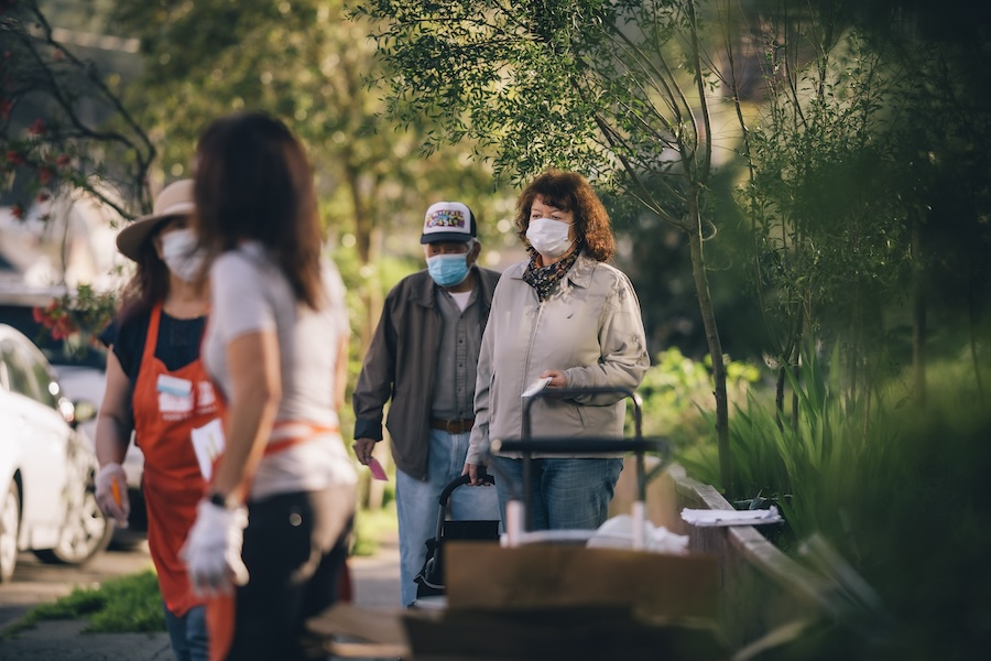 People wearing face masks walk along a sunny, plant-lined street with boxes nearby.