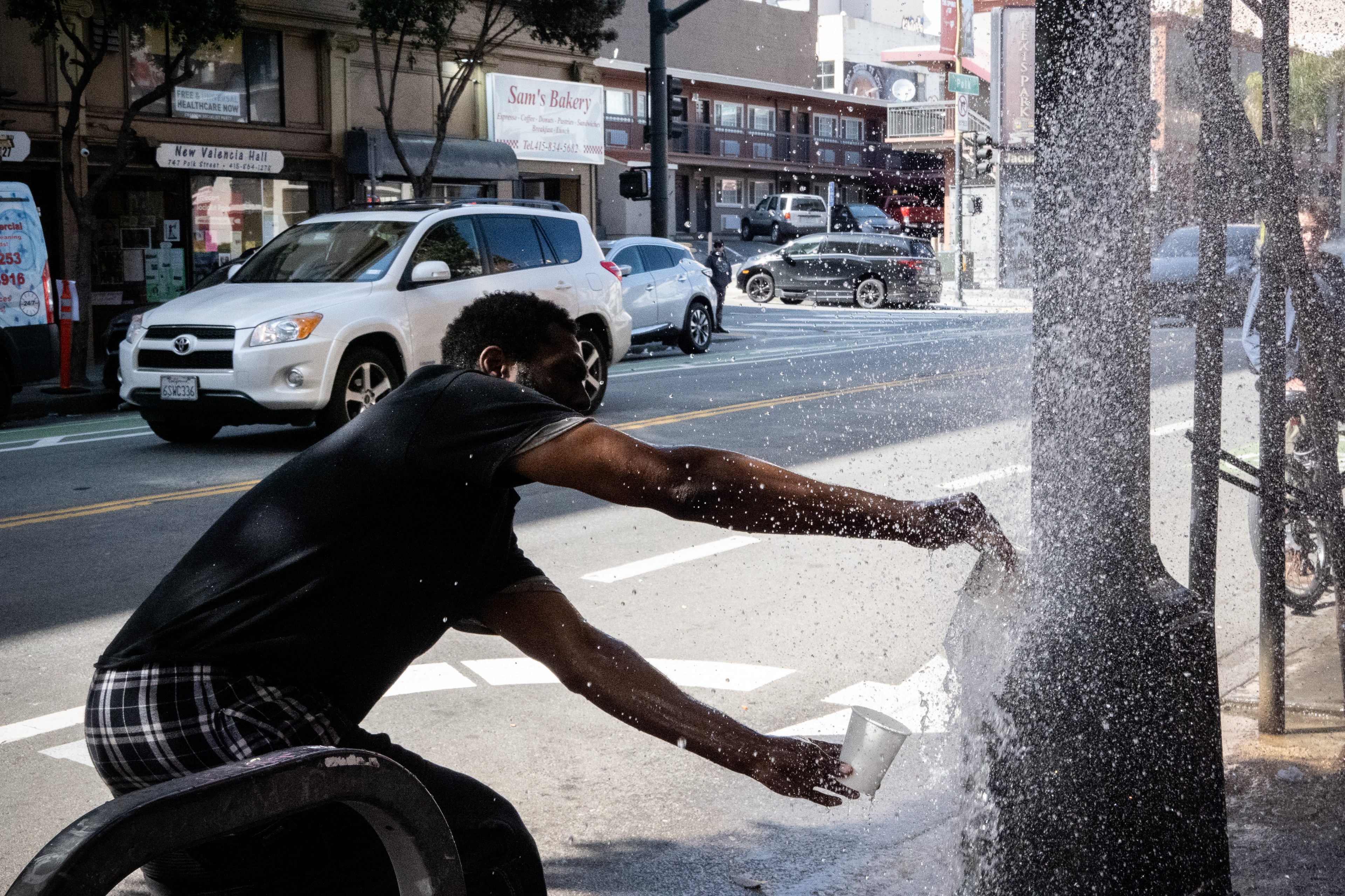 A person is splashing water from a street fountain, with vehicles and city backdrop.