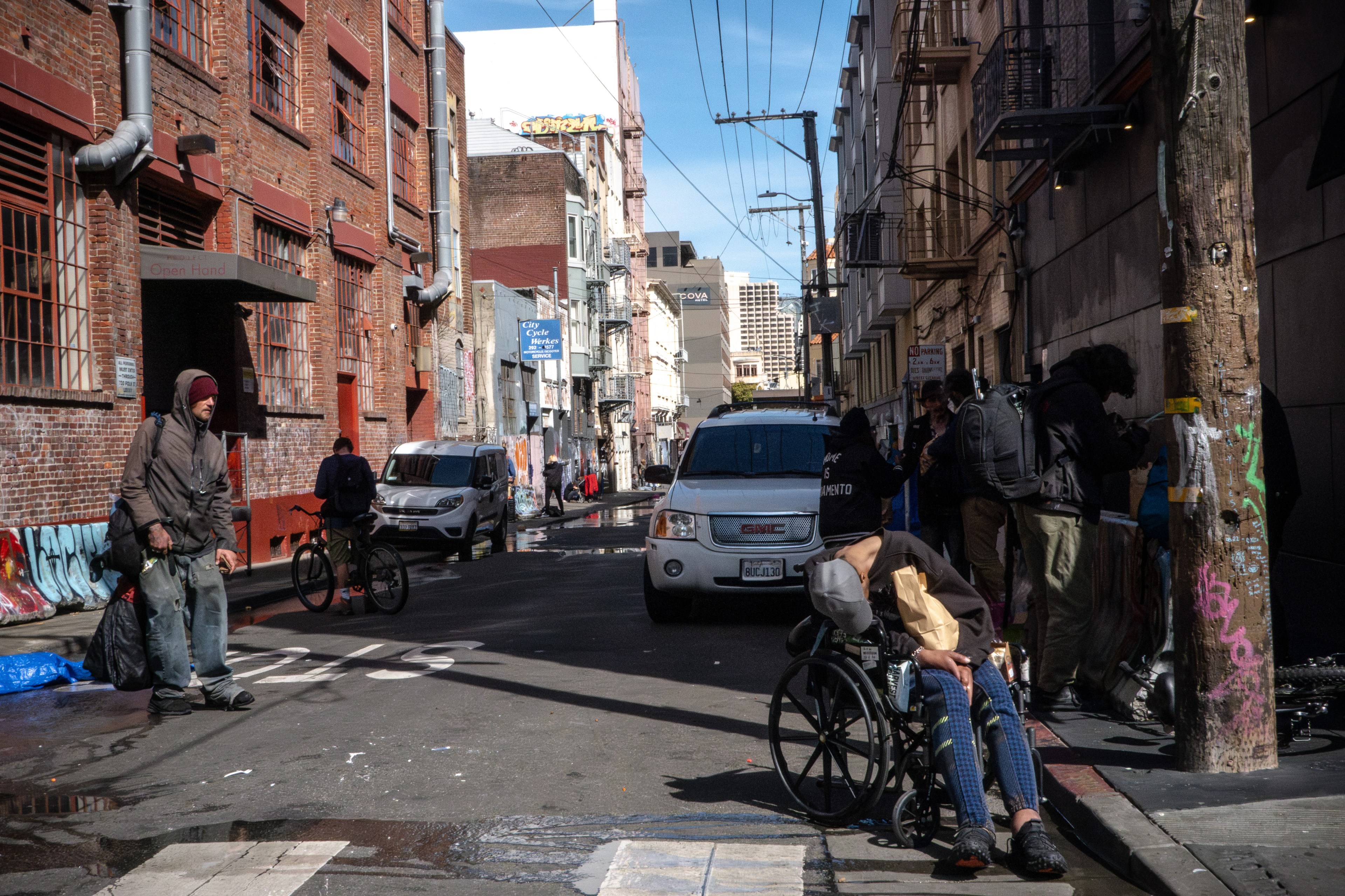 An urban alley with people, one in a wheelchair, graffiti, vehicles, and a cyclist in daylight.