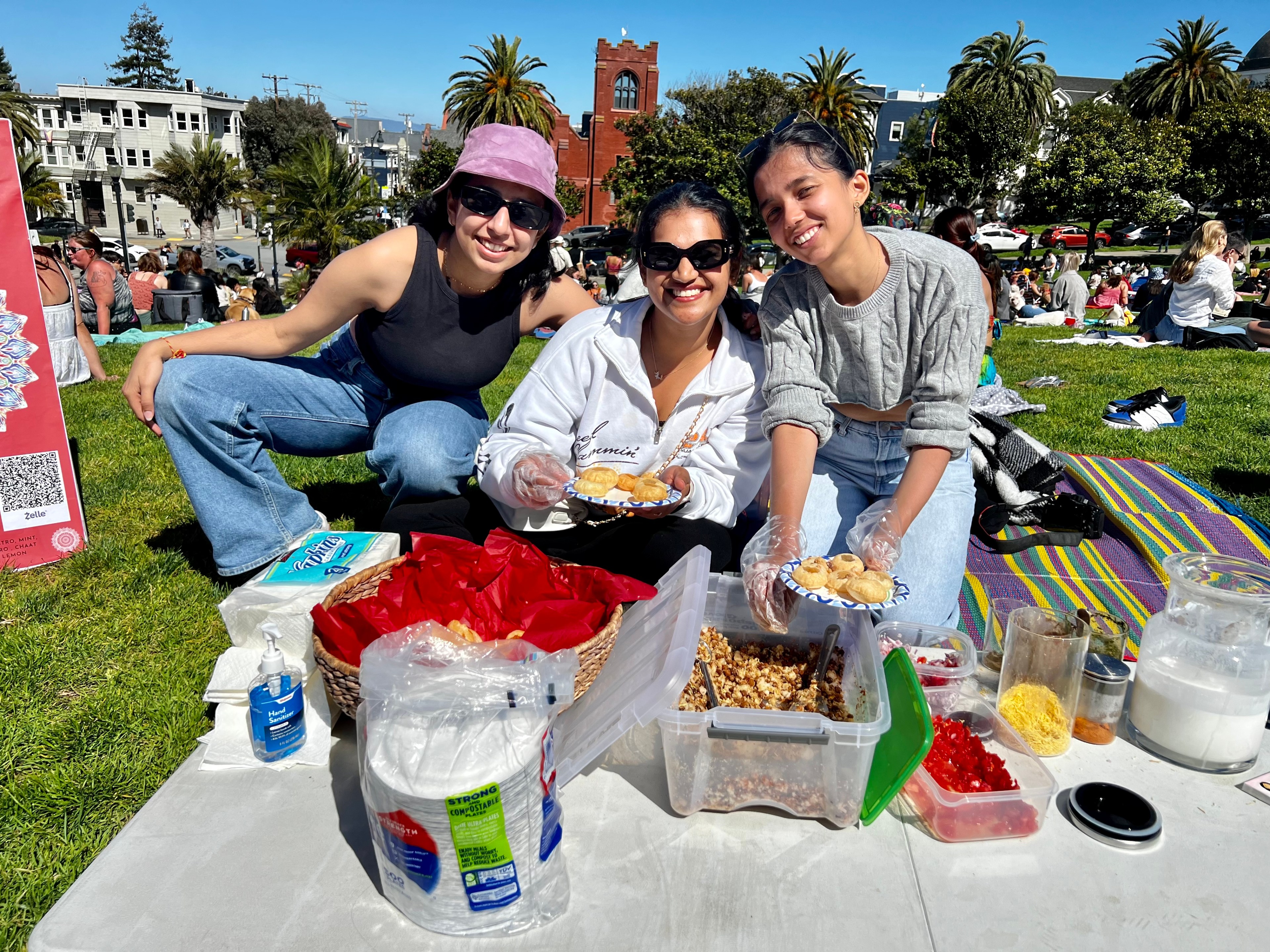 Three people sitting on a blanket with food at a sunny park, smiling at the camera.