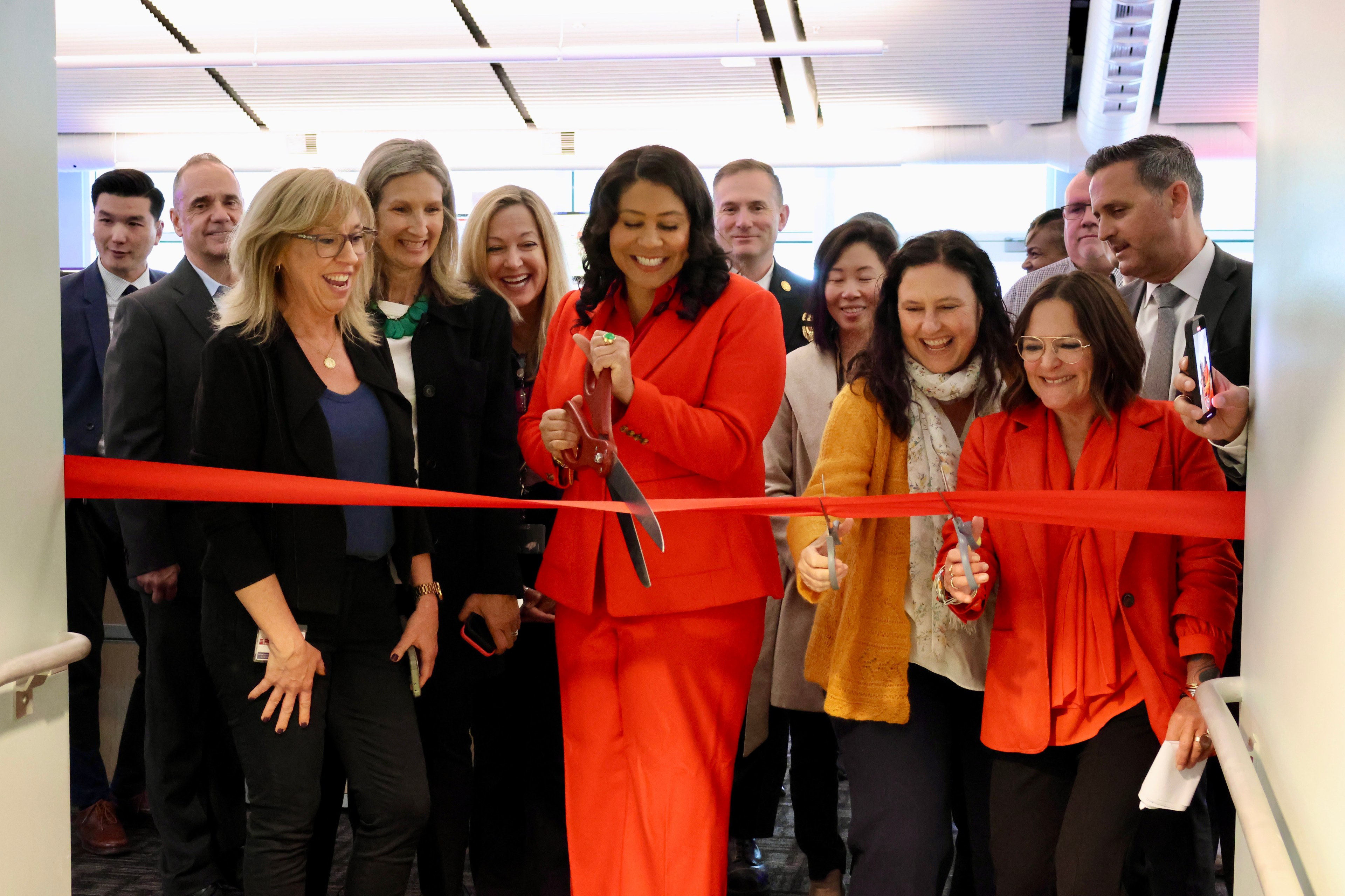 A group of smiling people watching a woman in a red suit cutting a ceremonial red ribbon.