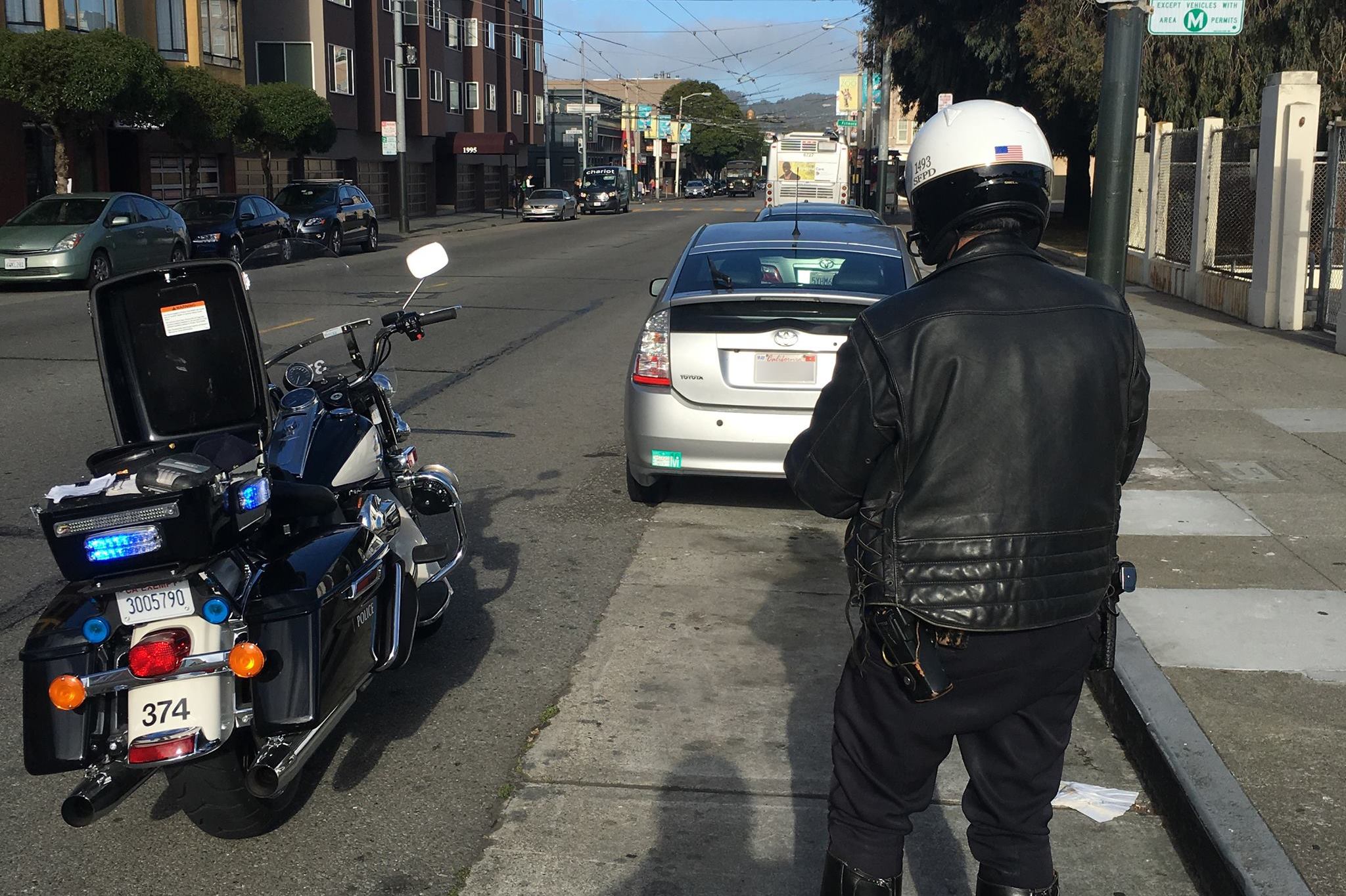 A police officer wearing a helmet and leather jacket is standing beside a motorcycle on a city street, writing a ticket for a parked silver Toyota Prius.