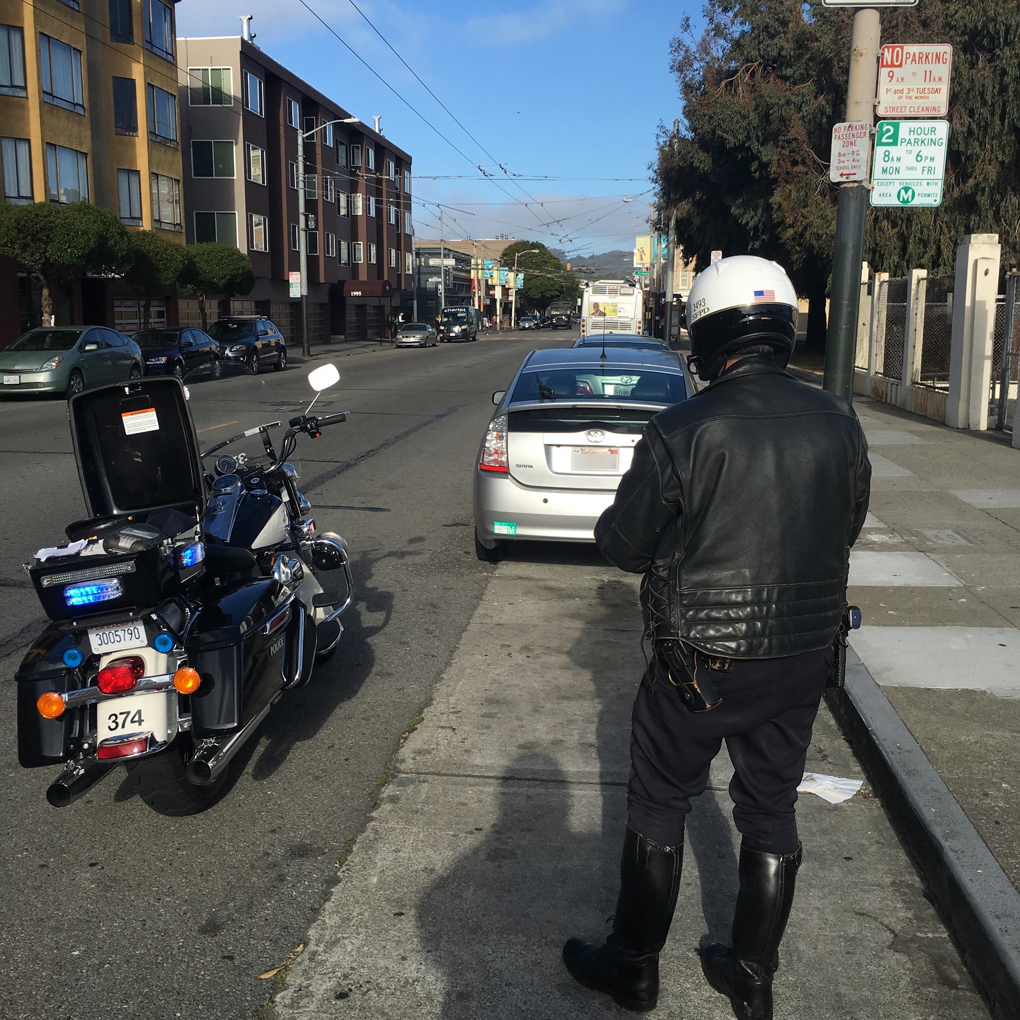 A police officer wearing a helmet and leather jacket is standing beside a motorcycle on a city street, writing a ticket for a parked silver Toyota Prius.