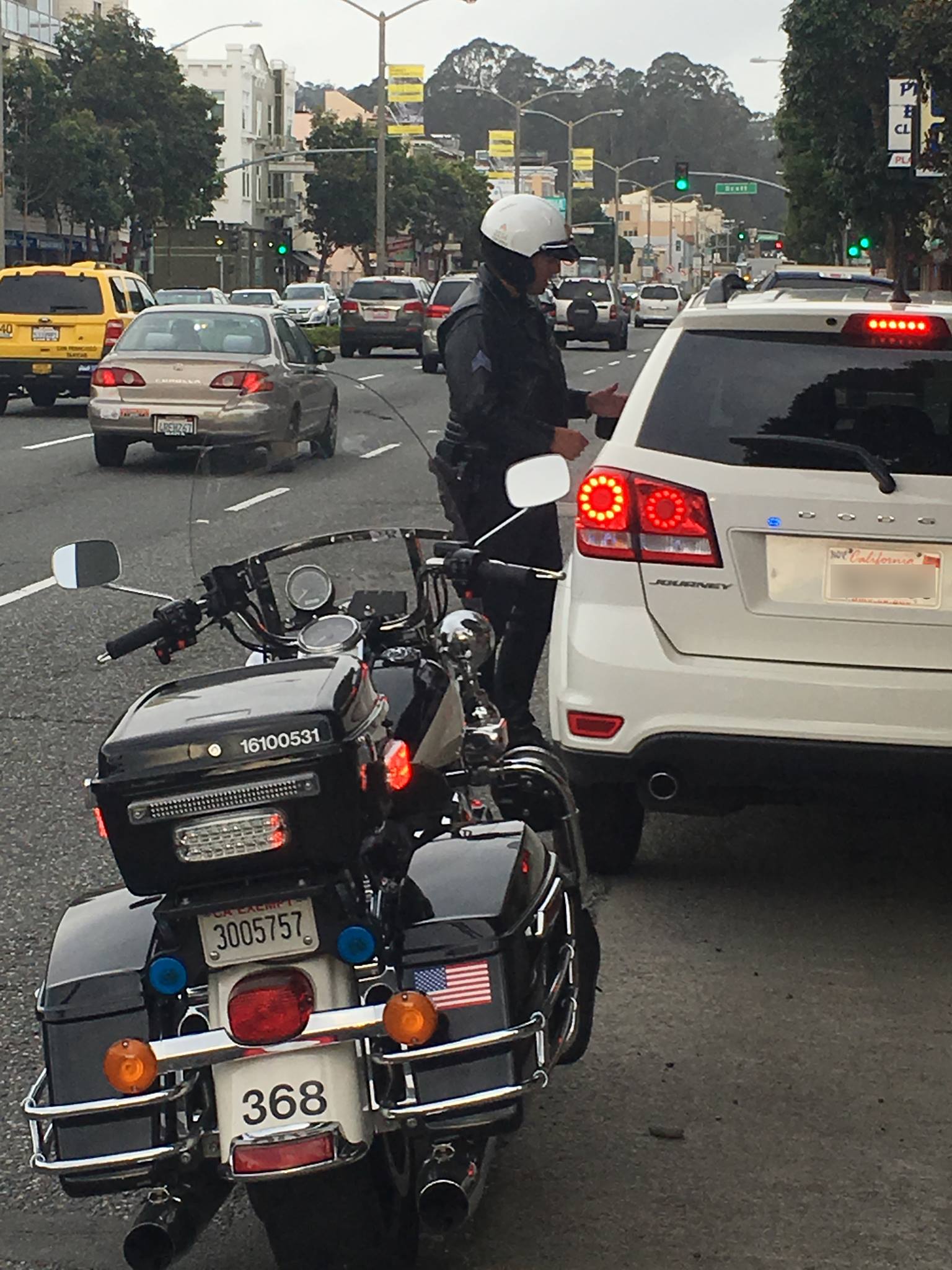 A police officer on a motorcycle is stopped beside a white car on a busy street.