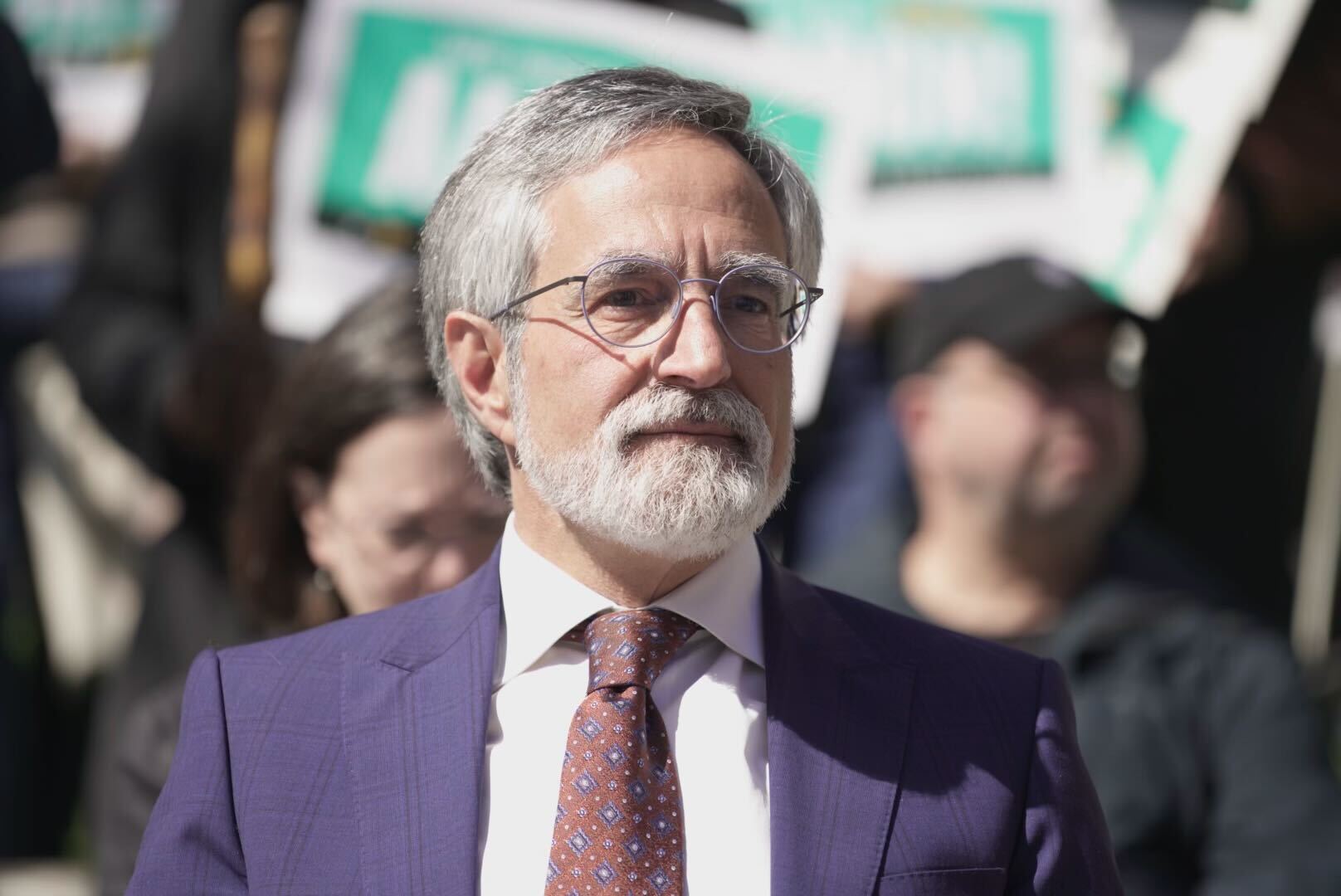 A man with gray hair and a beard, wearing glasses, a purple suit, and a patterned tie.