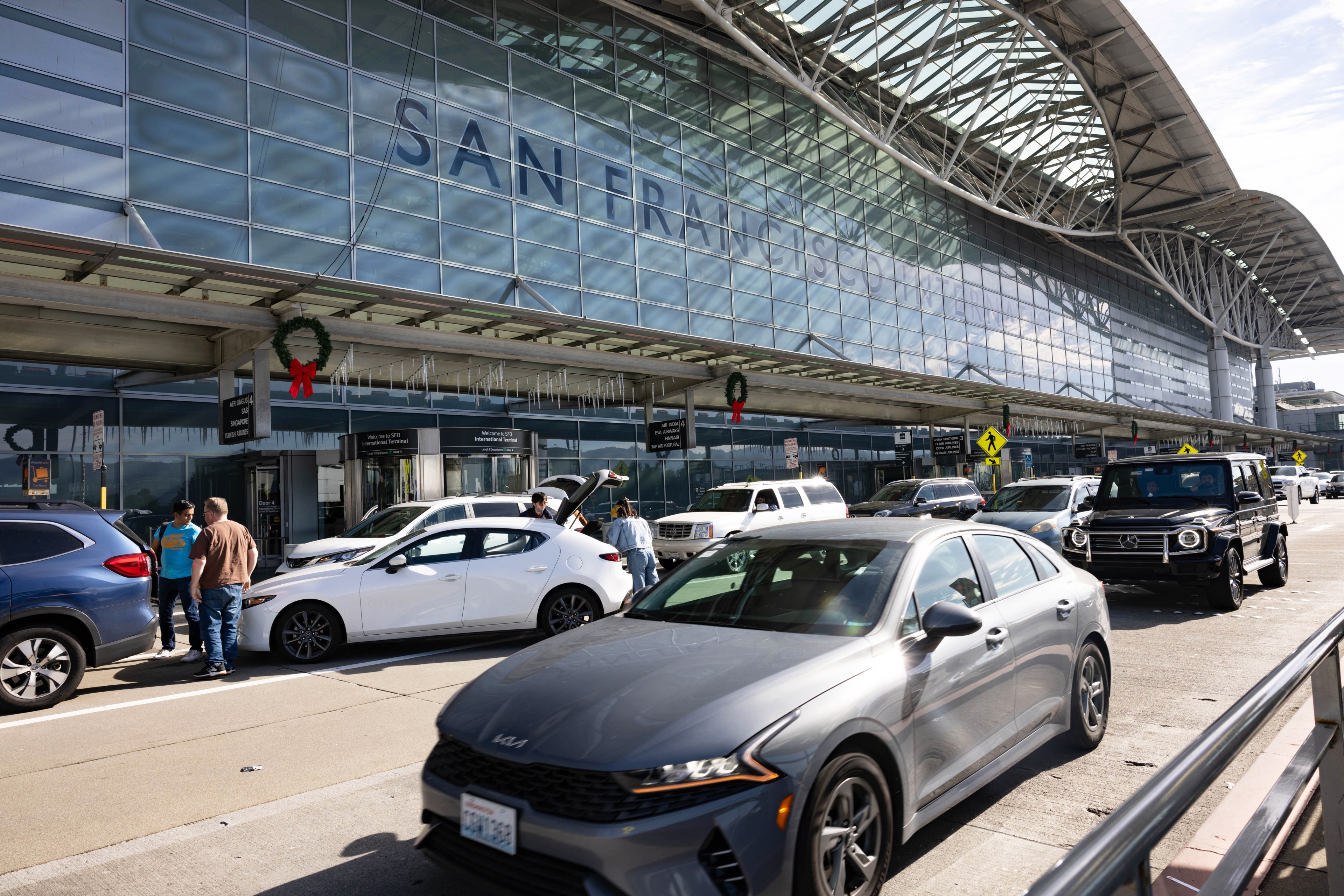 The image shows a busy airport terminal with cars lined up for drop-offs. People are loading and unloading luggage. Holiday wreaths decorate the facade.