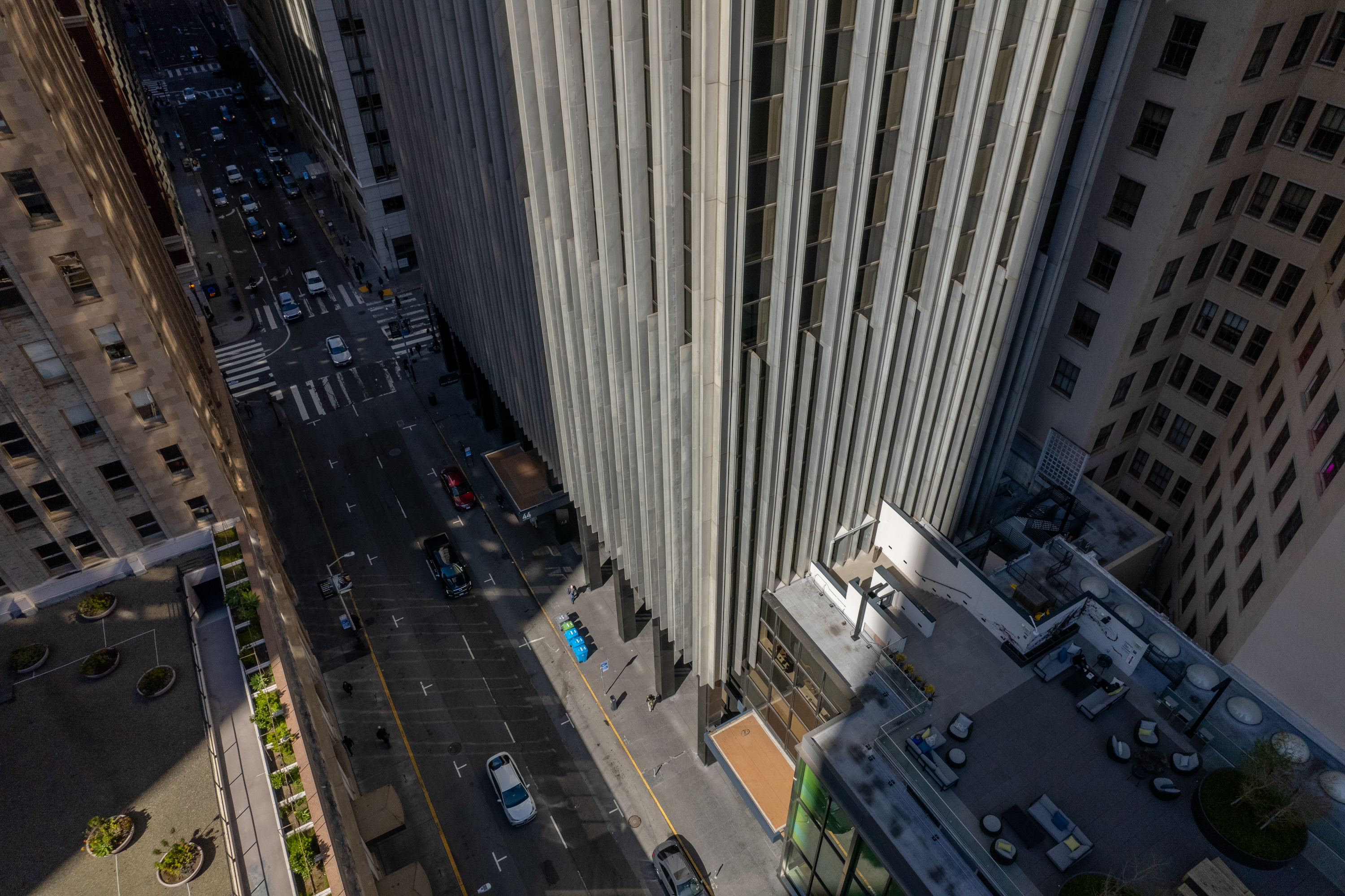 Aerial view of city street between tall buildings, casting shadows, with visible cars and a rooftop patio.