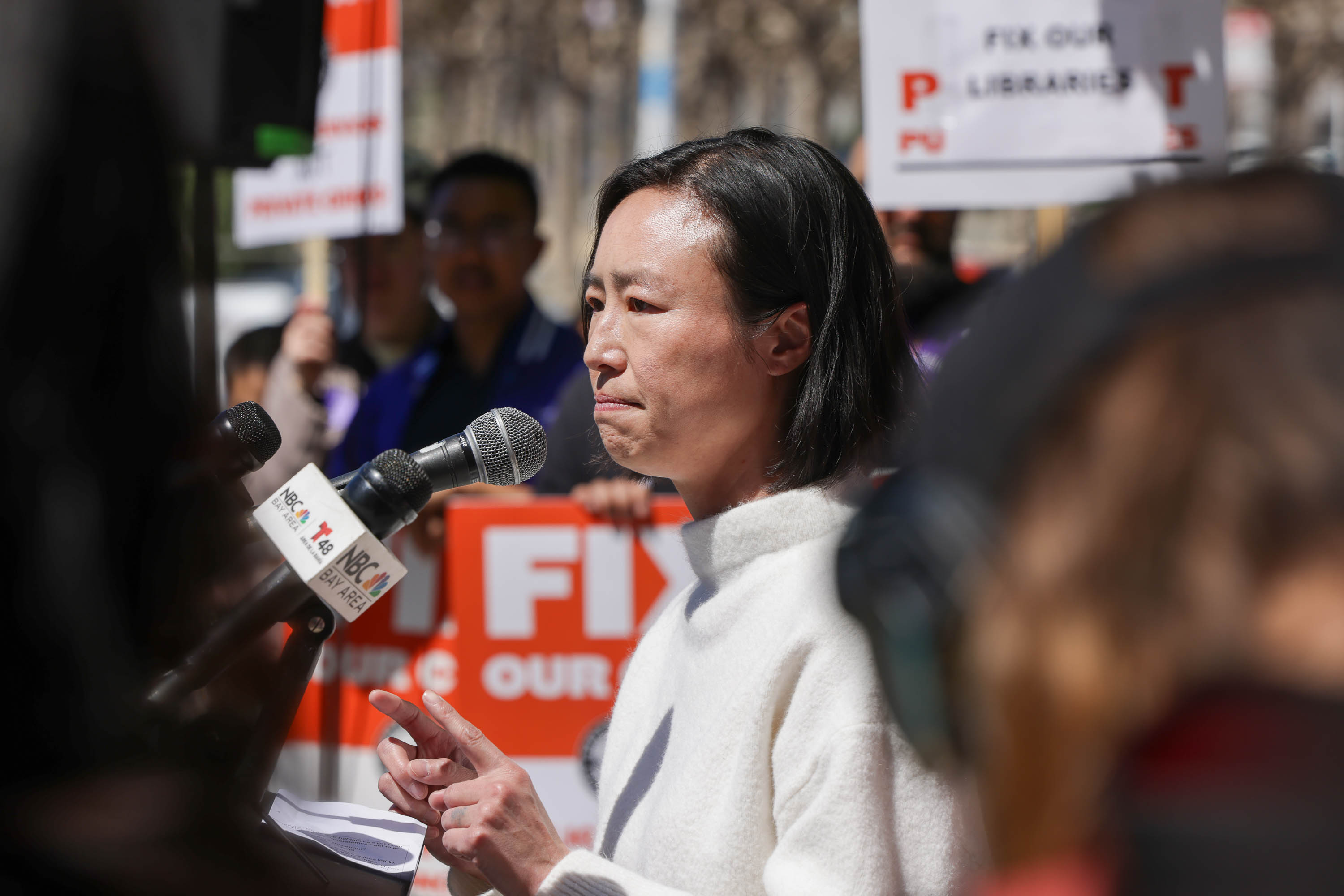 A woman in a white sweater speaks into microphones during a protest. People holding &quot;Fix Our Libraries&quot; signs are visible in the background.