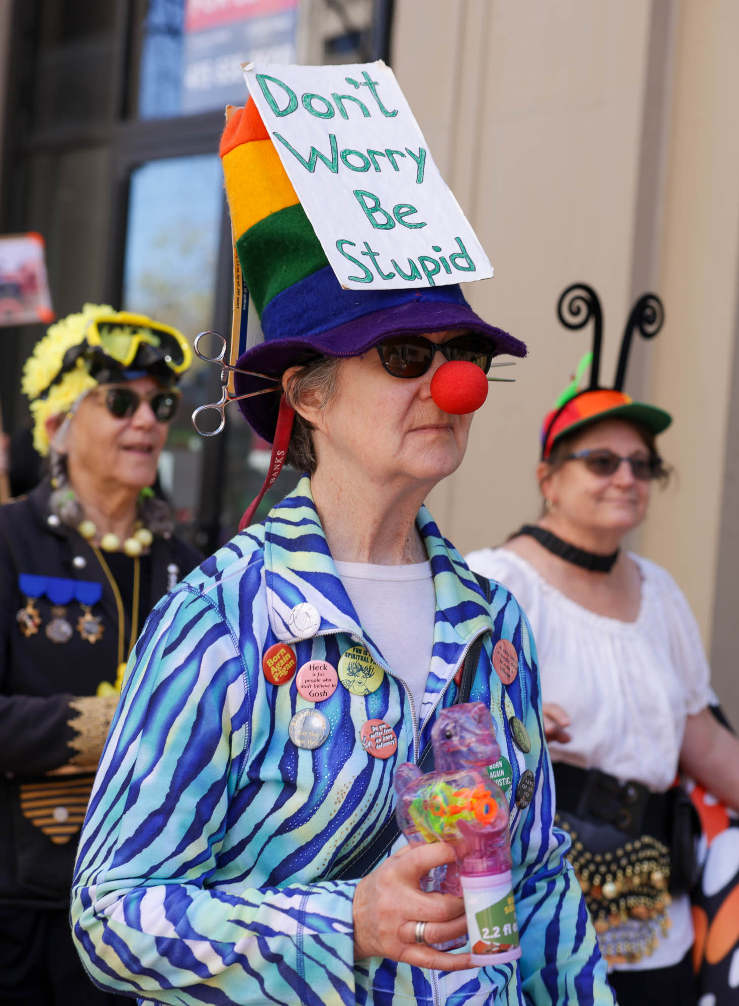 A person in a colorful hat with a &quot;Don't Worry Be Stupid&quot; sign, red nose, and a button-adorned jacket is holding a quirky drink cup.