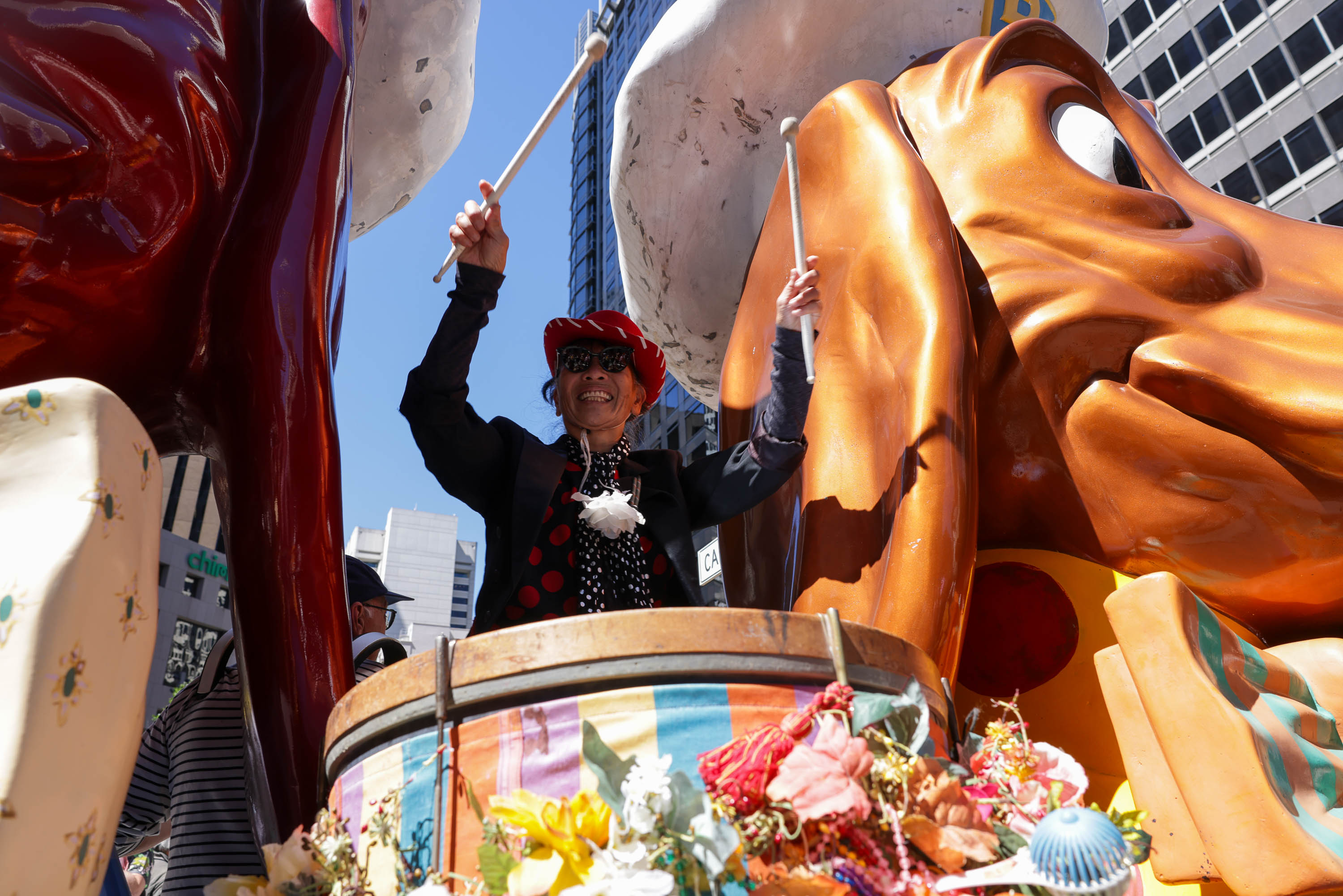 A person in a red hat and polka-dot outfit plays drums on a colorful float with large chicken sculptures towering behind.