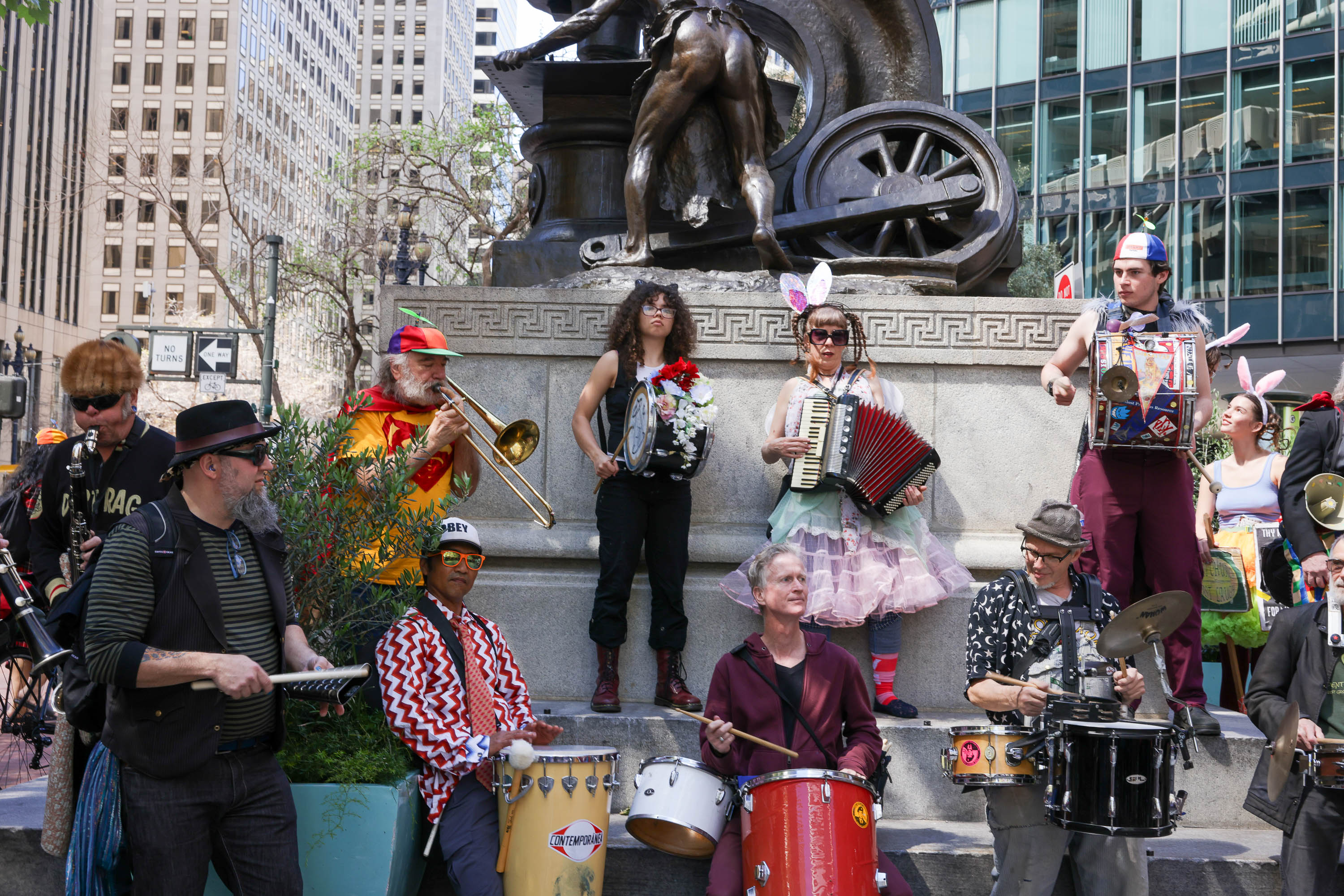 A lively street band with eclectic outfits plays instruments near a statue in an urban setting.