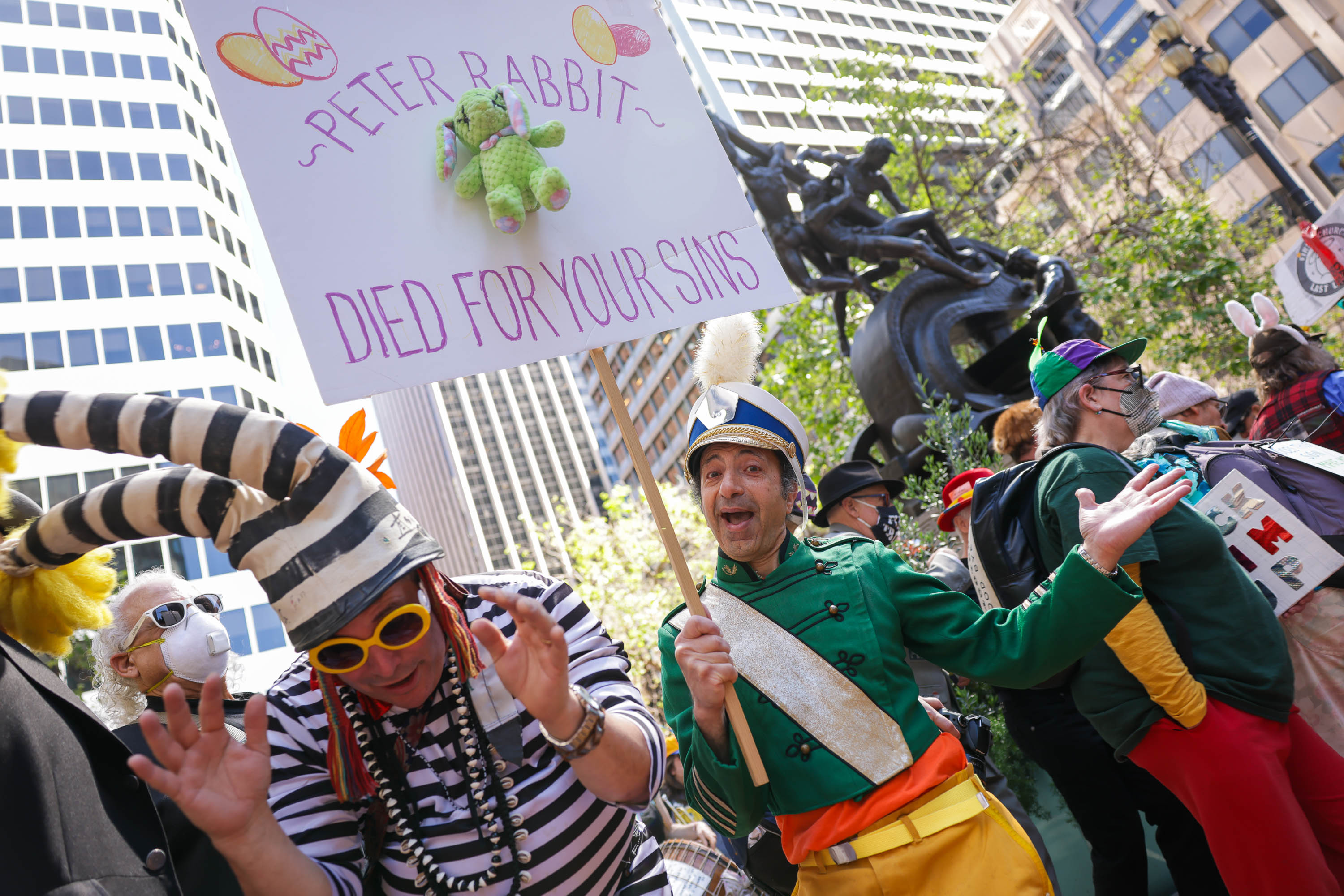 People in colorful costumes, one holding a sign saying &quot;Peter Rabbit died for your sins,&quot; at an outdoor gathering.