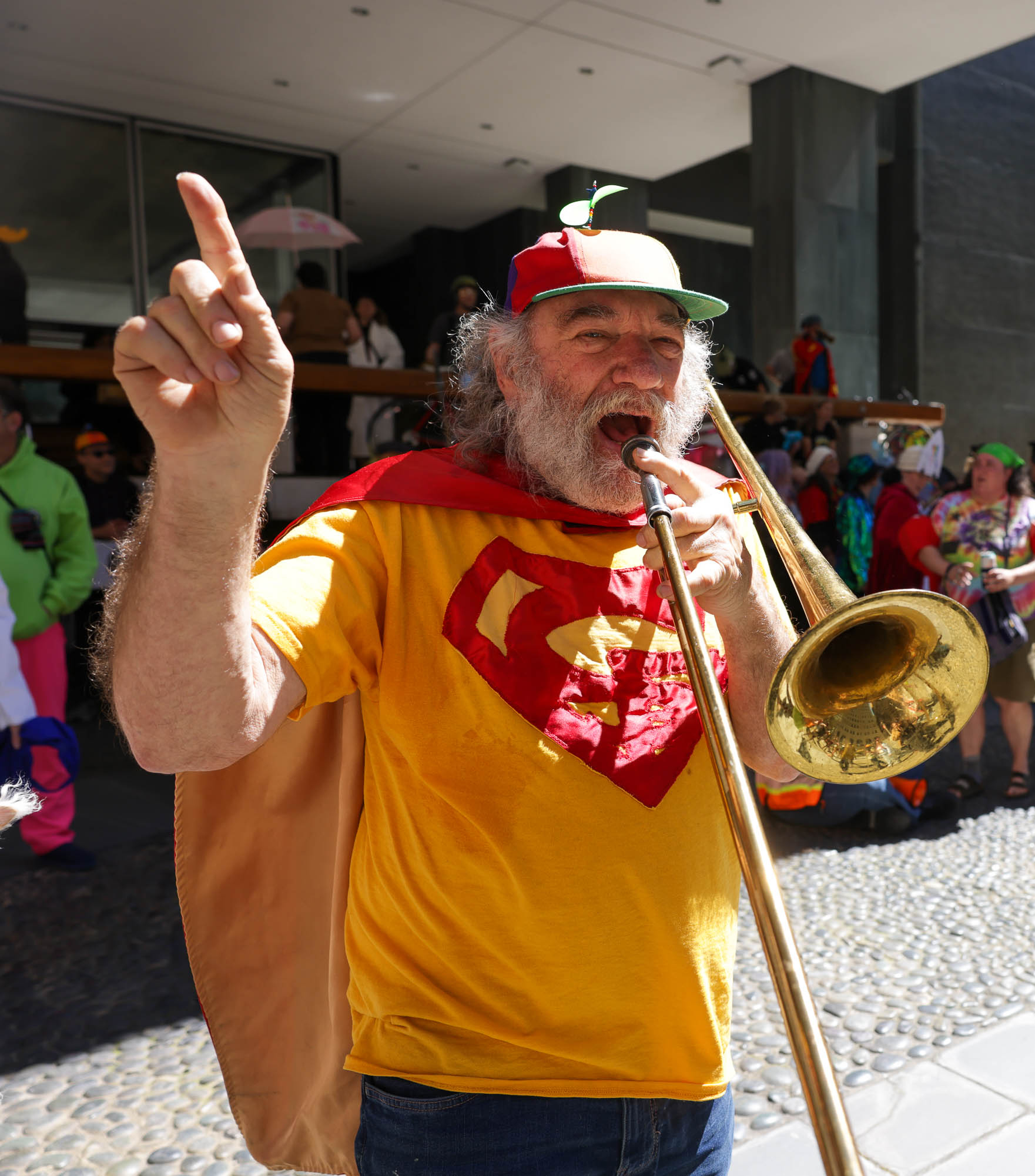 A man in a Superman shirt and hat plays a trombone, pointing skyward, at an outdoor event.