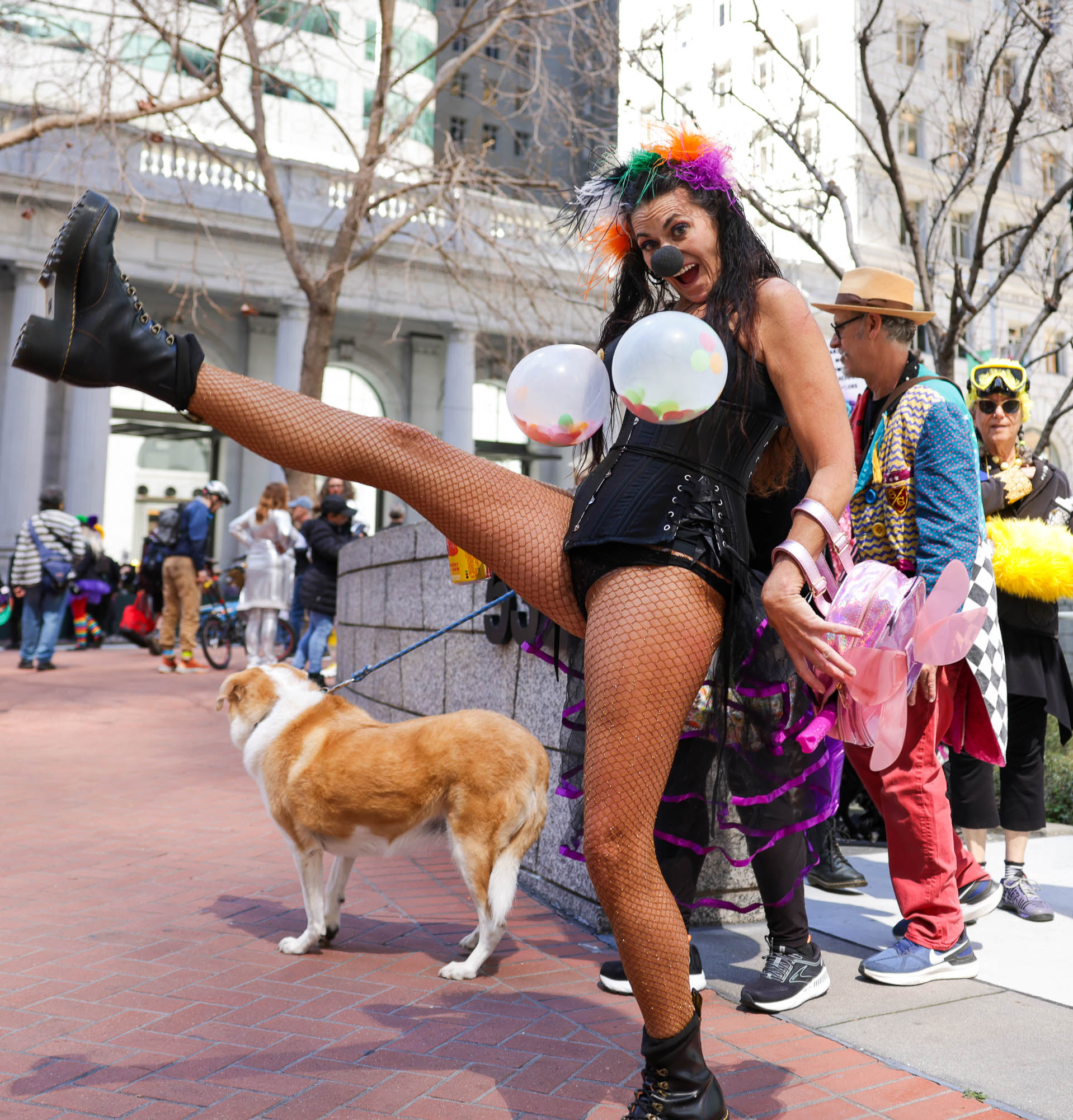 A person in colorful costume and makeup raises a leg high, fishnets visible, with a dog and people in festive wear behind.