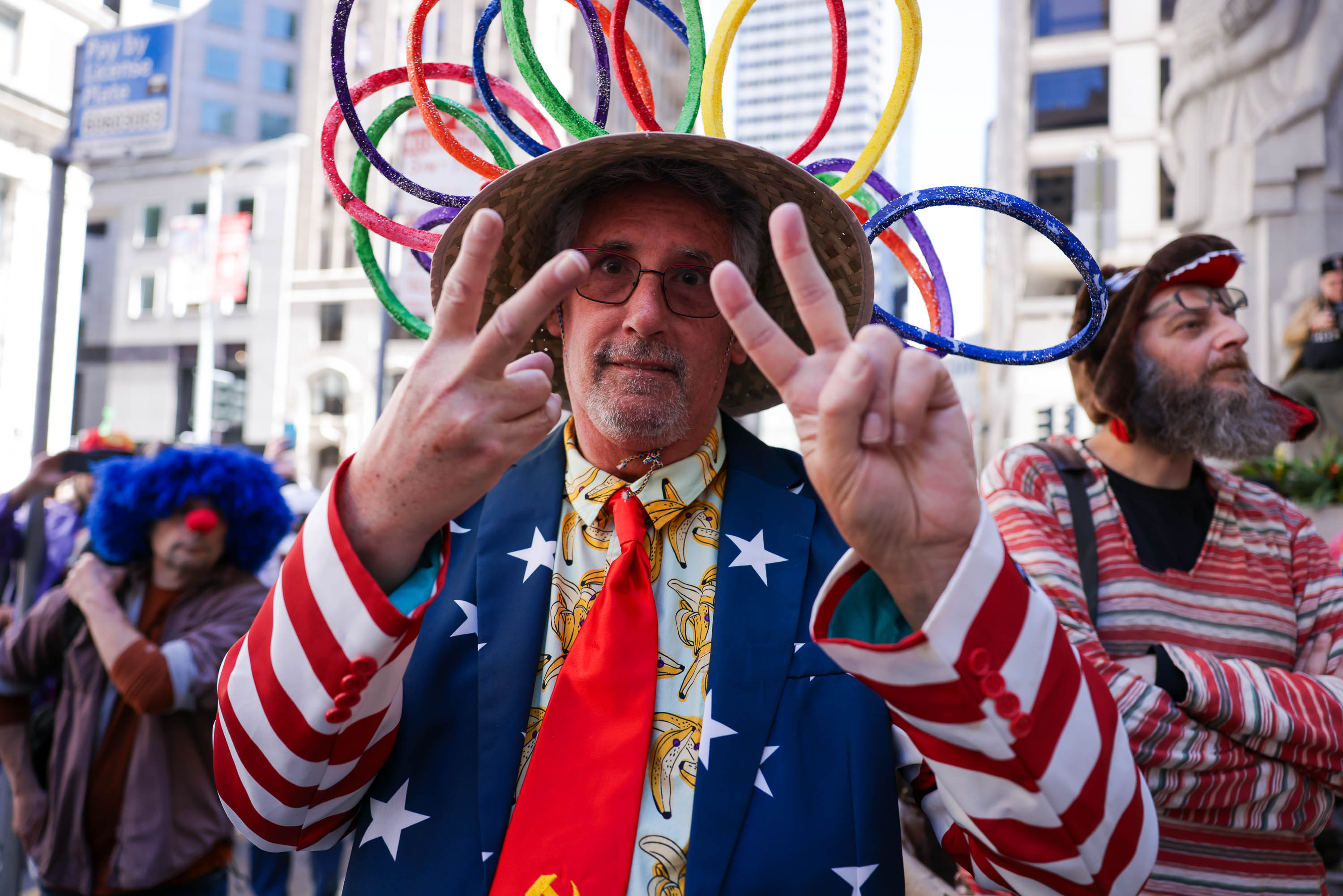A man in a colorful hat and star-spangled jacket gesturing a peace sign, with a clown in the background.