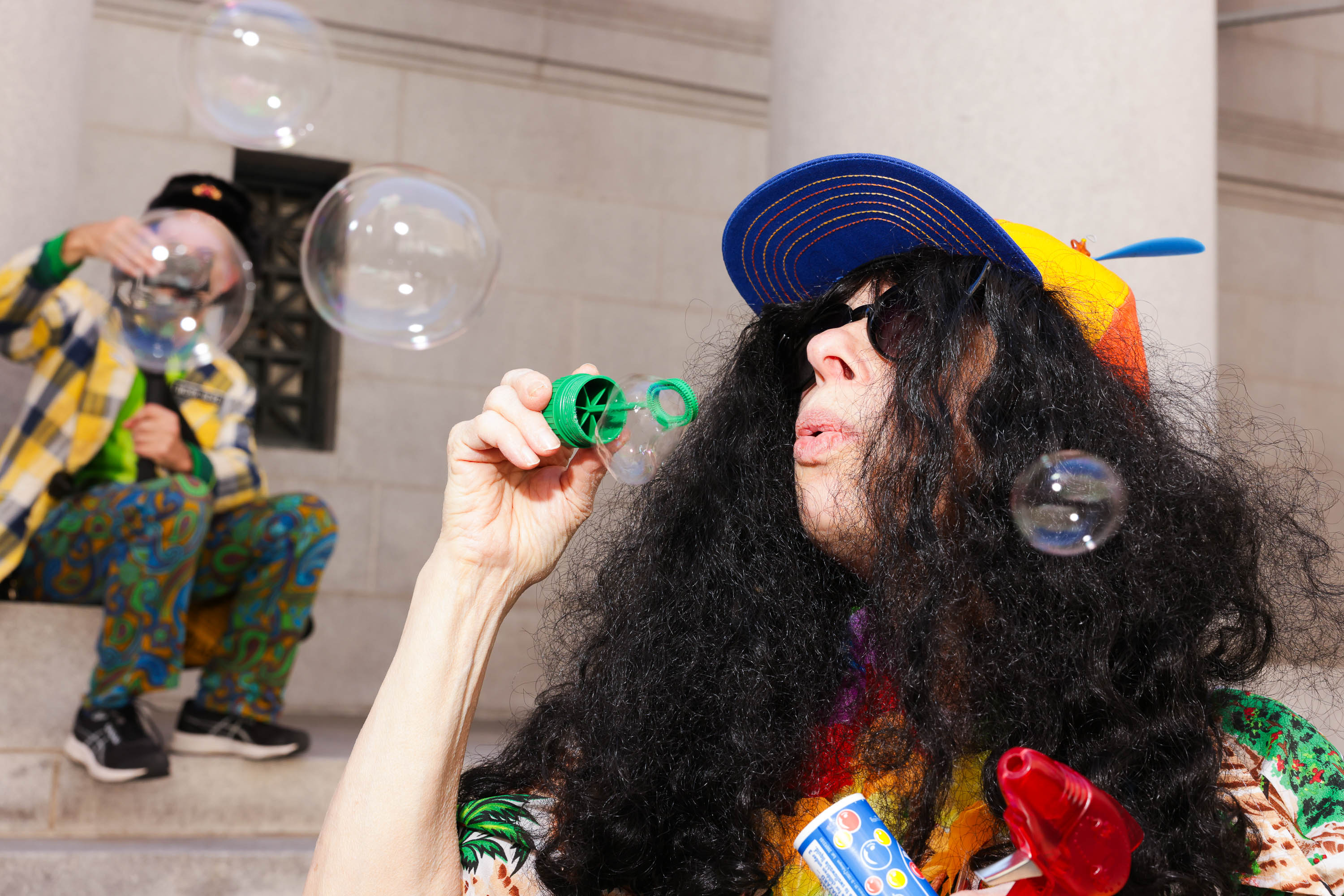 Two people in colorful costumes blow bubbles; one in foreground is wearing sunglasses and a multicolored cap.