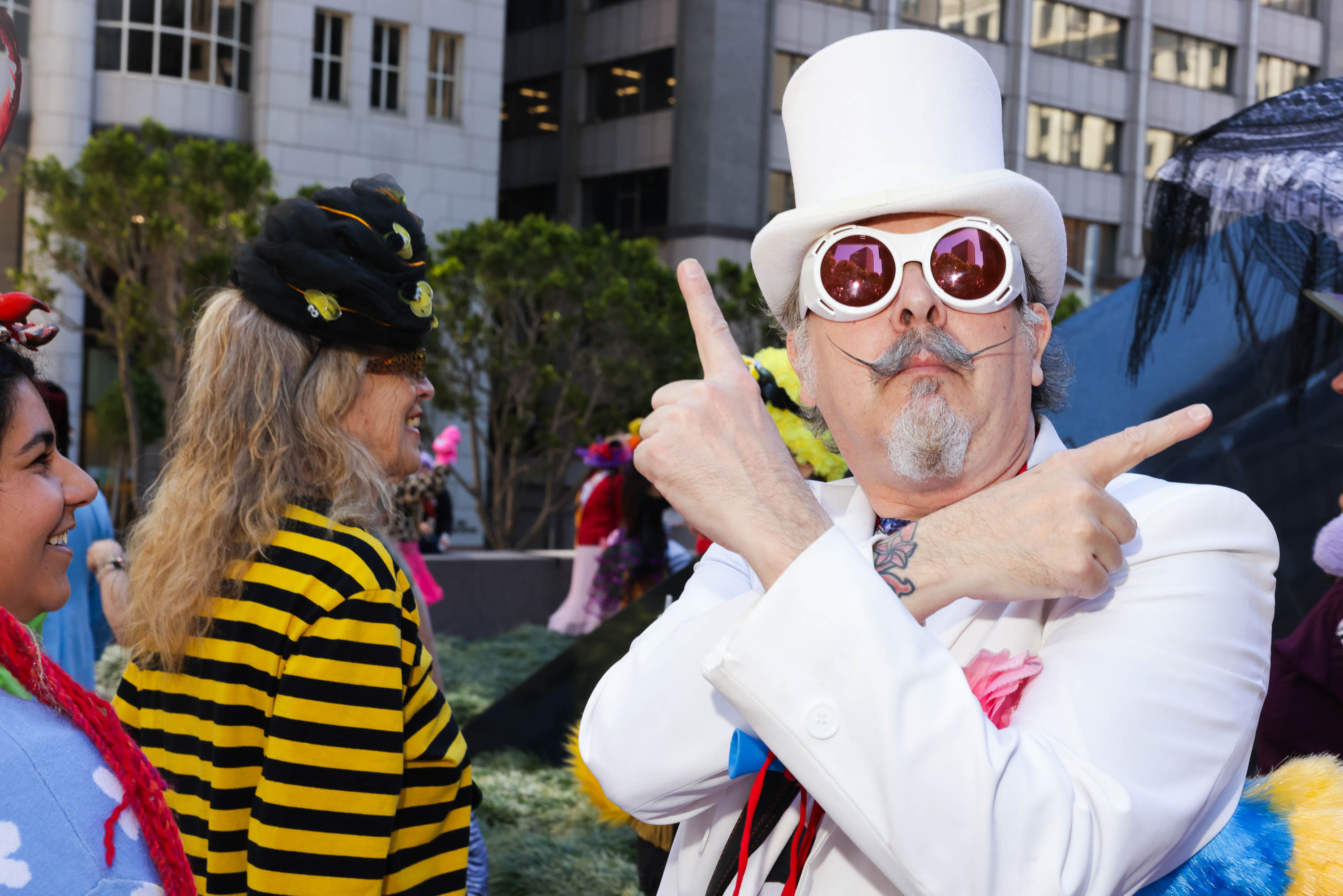 A man in a white top hat and sunglasses strikes a playful pose with two women in colorful, whimsical outfits.