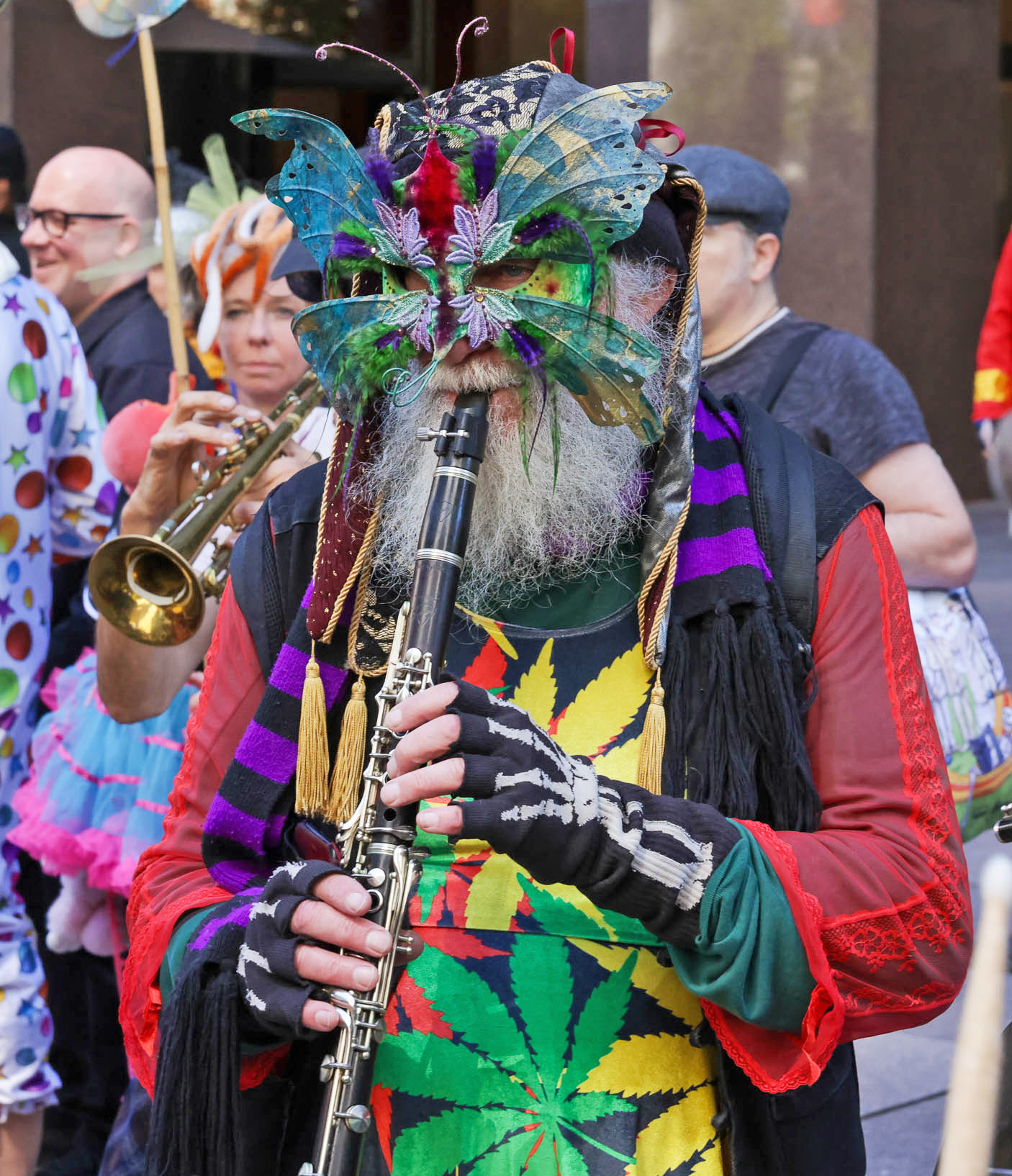 A person in a vibrant butterfly mask and colorful attire plays the clarinet in a street parade.