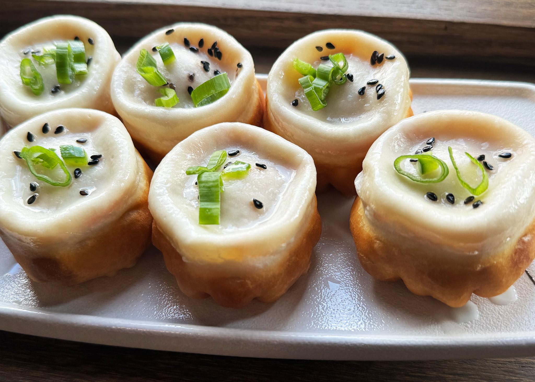 Six round, translucent dumplings topped with green onions and black sesame seeds on a plate.