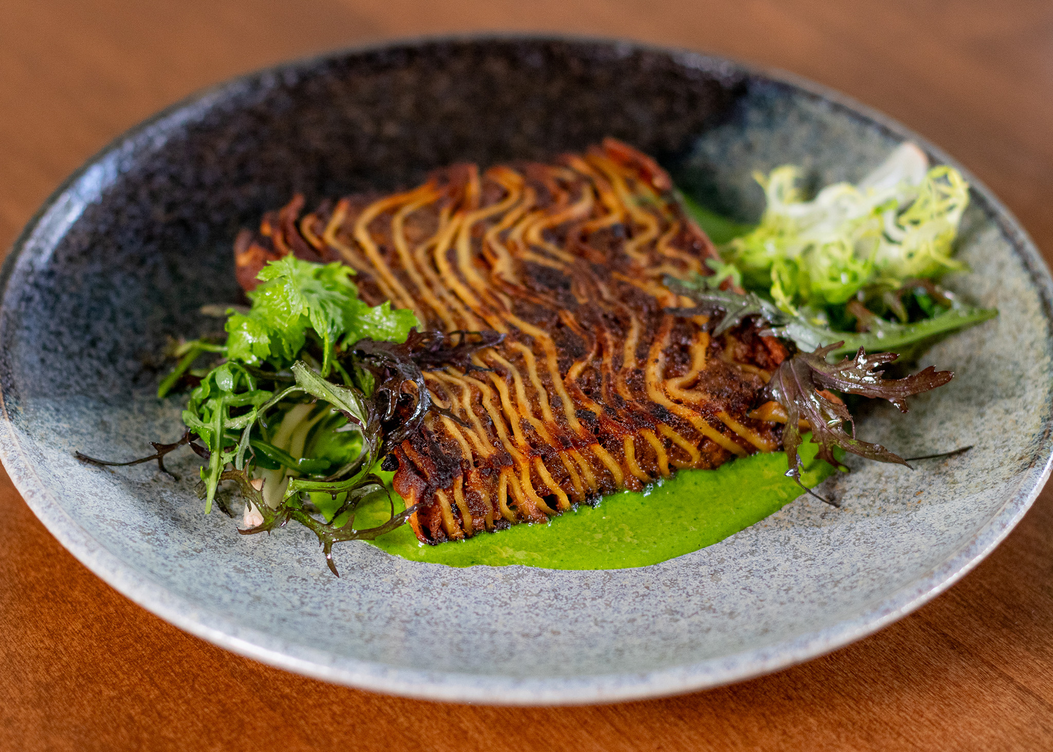 Grilled fish with charred top on a bed of green sauce, garnished with leafy greens, served on a speckled blue plate.