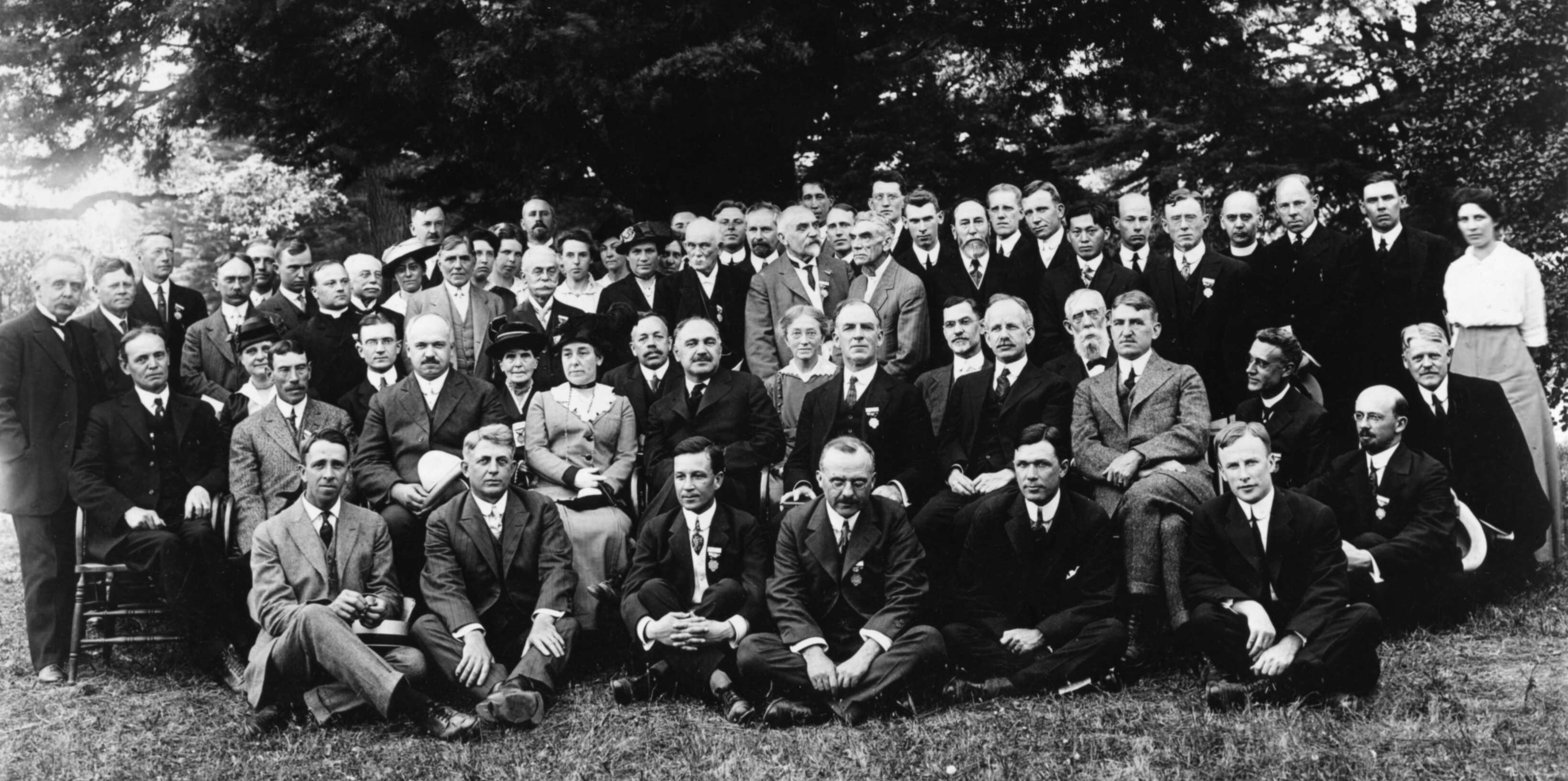 A group of formally dressed people, mostly men with a few women, posing outdoors for a historical group photo.