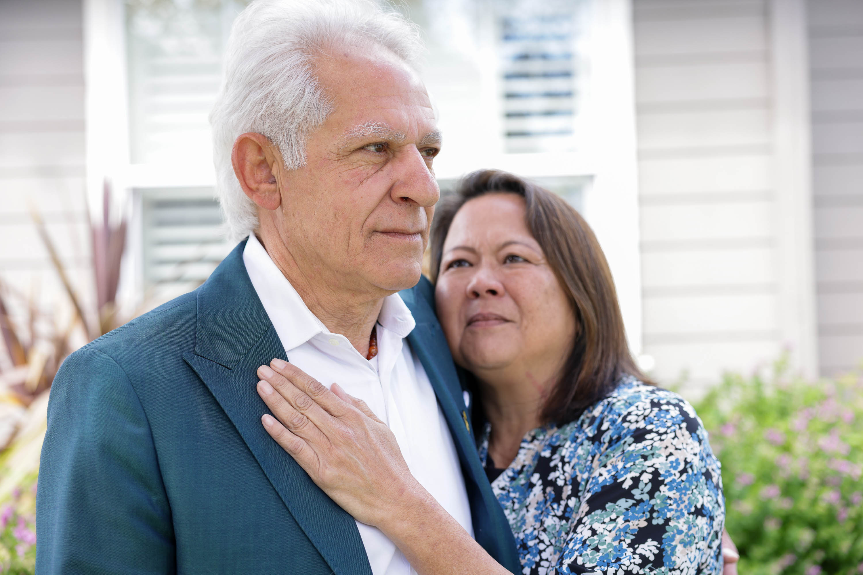 An older couple stands close, the woman embracing the man from behind, both looking thoughtful, in daylight. 