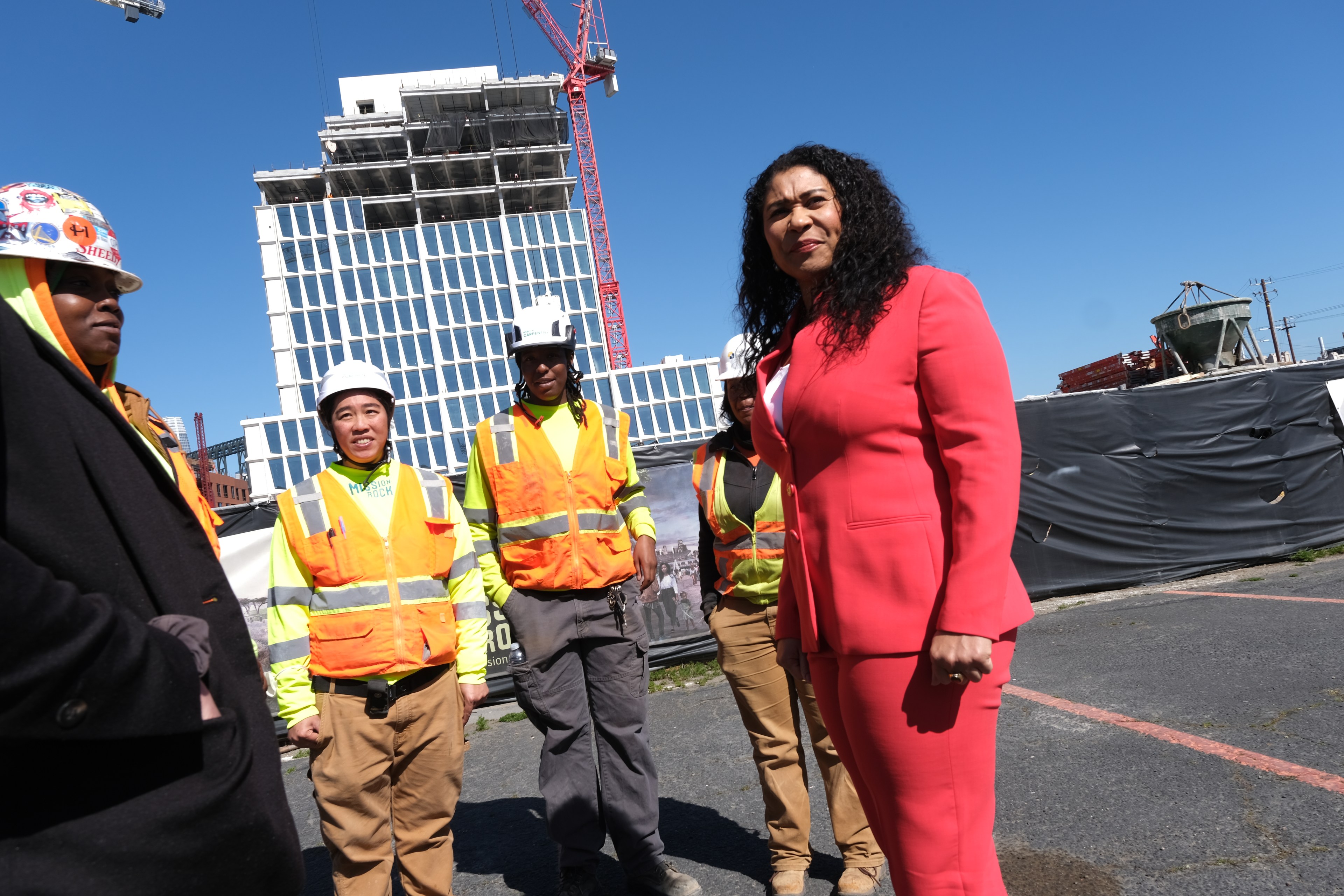 A woman in a red suit stands in the foreground with construction workers in high-visibility vests and a building site behind them.