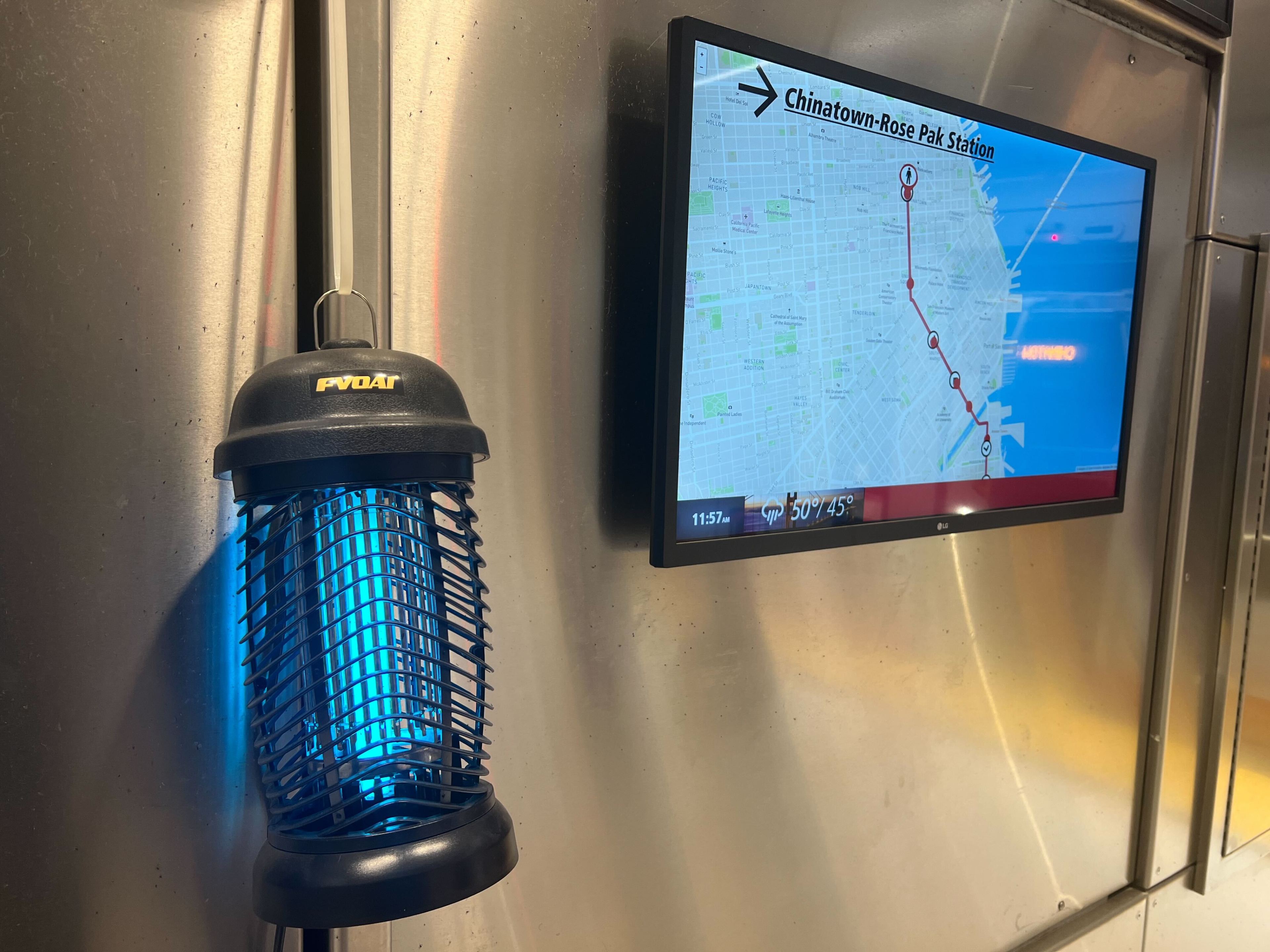 An indoor bug zapper hangs beside a digital screen displaying a transit map and the time/temp.