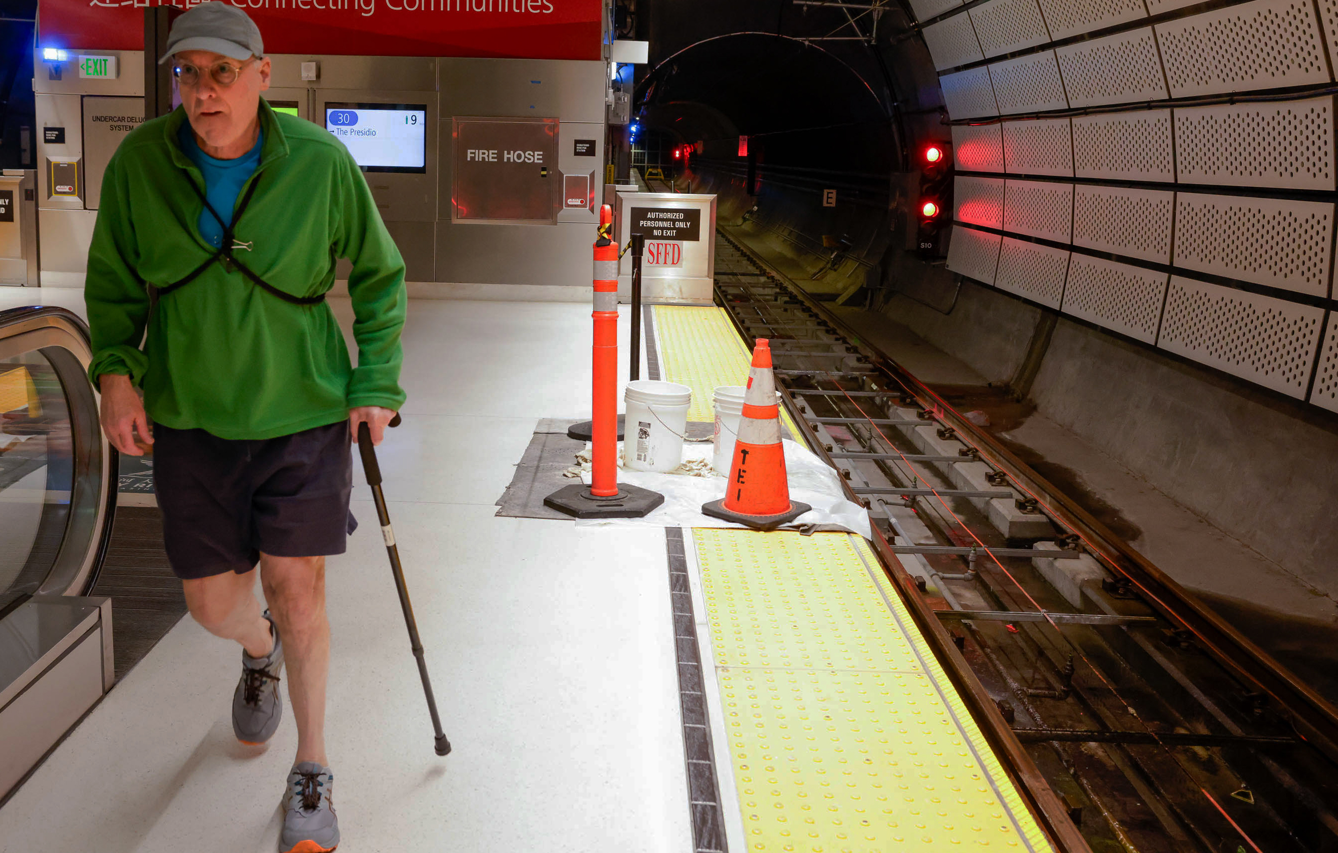 A man with a cane walks by subway tracks with traffic cones and construction signs.