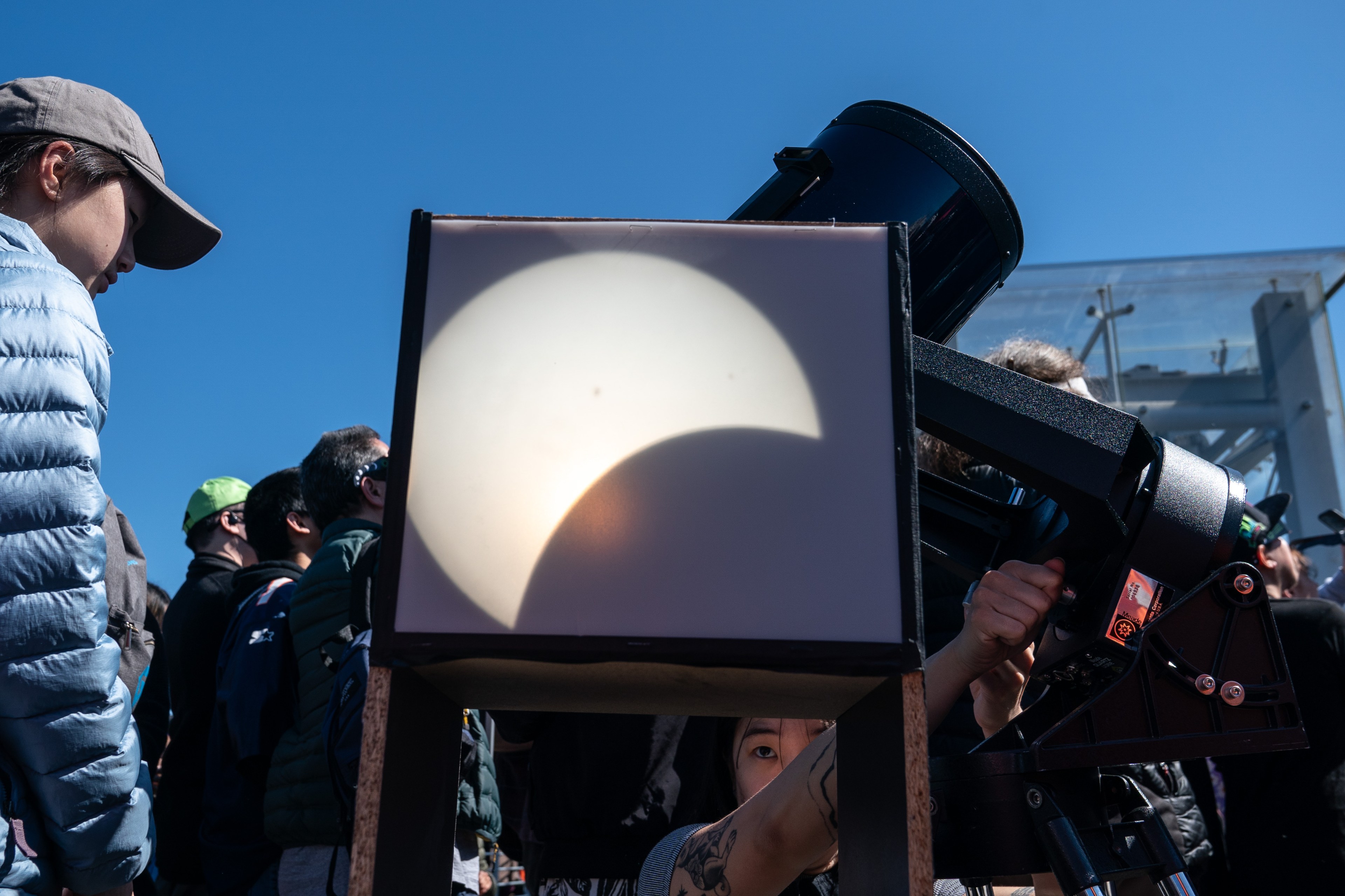 A solar eclipse projection on a screen attached to a telescope, with onlookers observing the event.