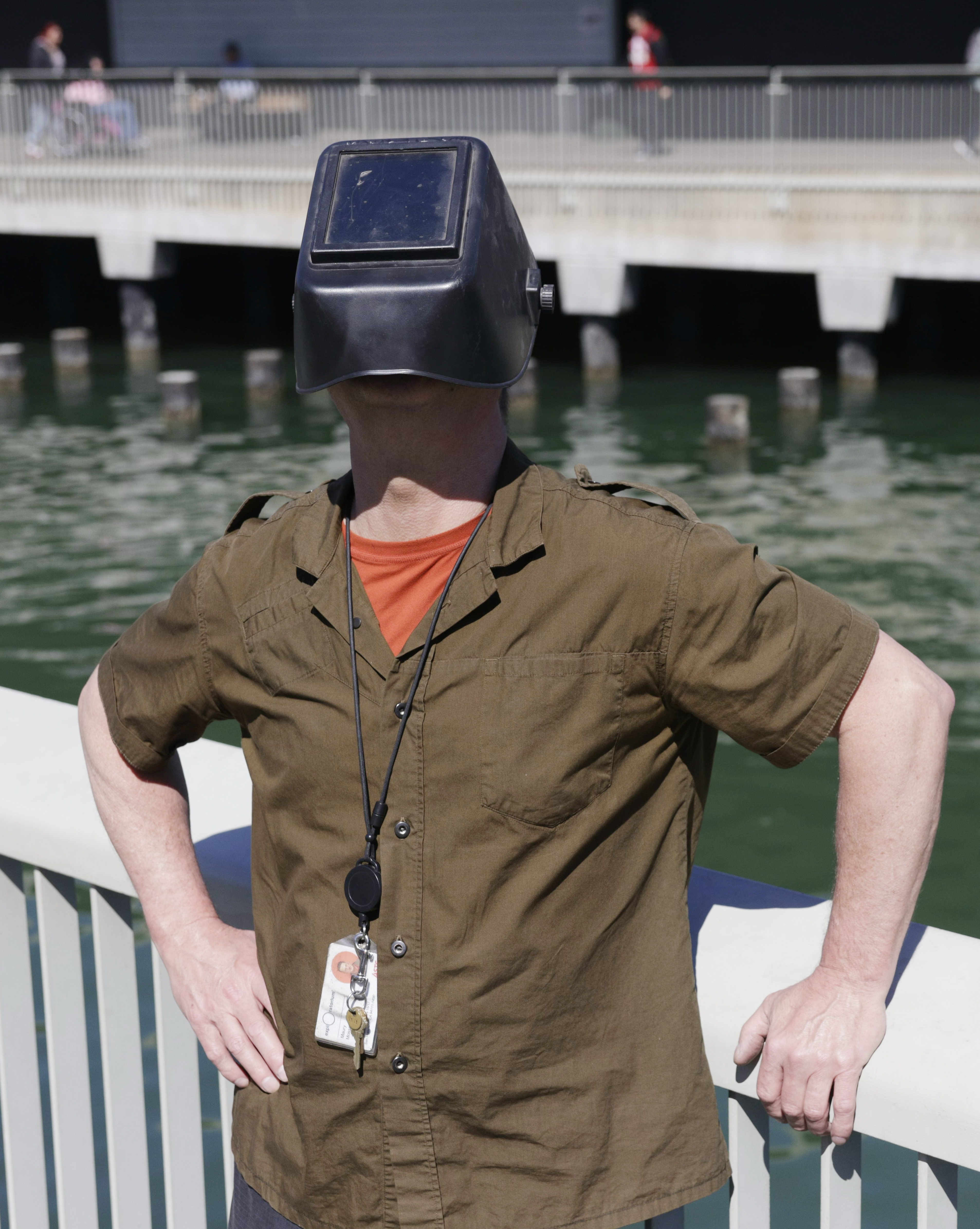 A person is standing by water, wearing a welding mask, khaki shirt, and an ID badge.
