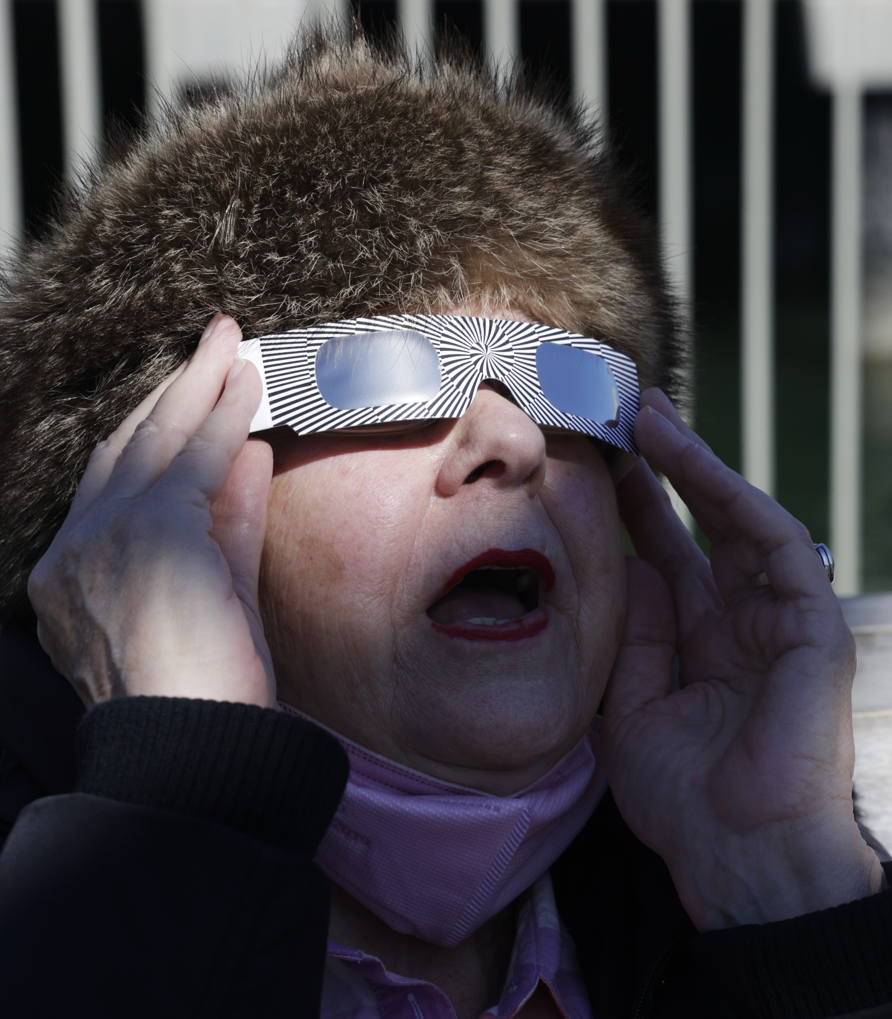 A person is wearing special glasses, likely viewing an eclipse, with a fur hat and an open mouth in awe.
