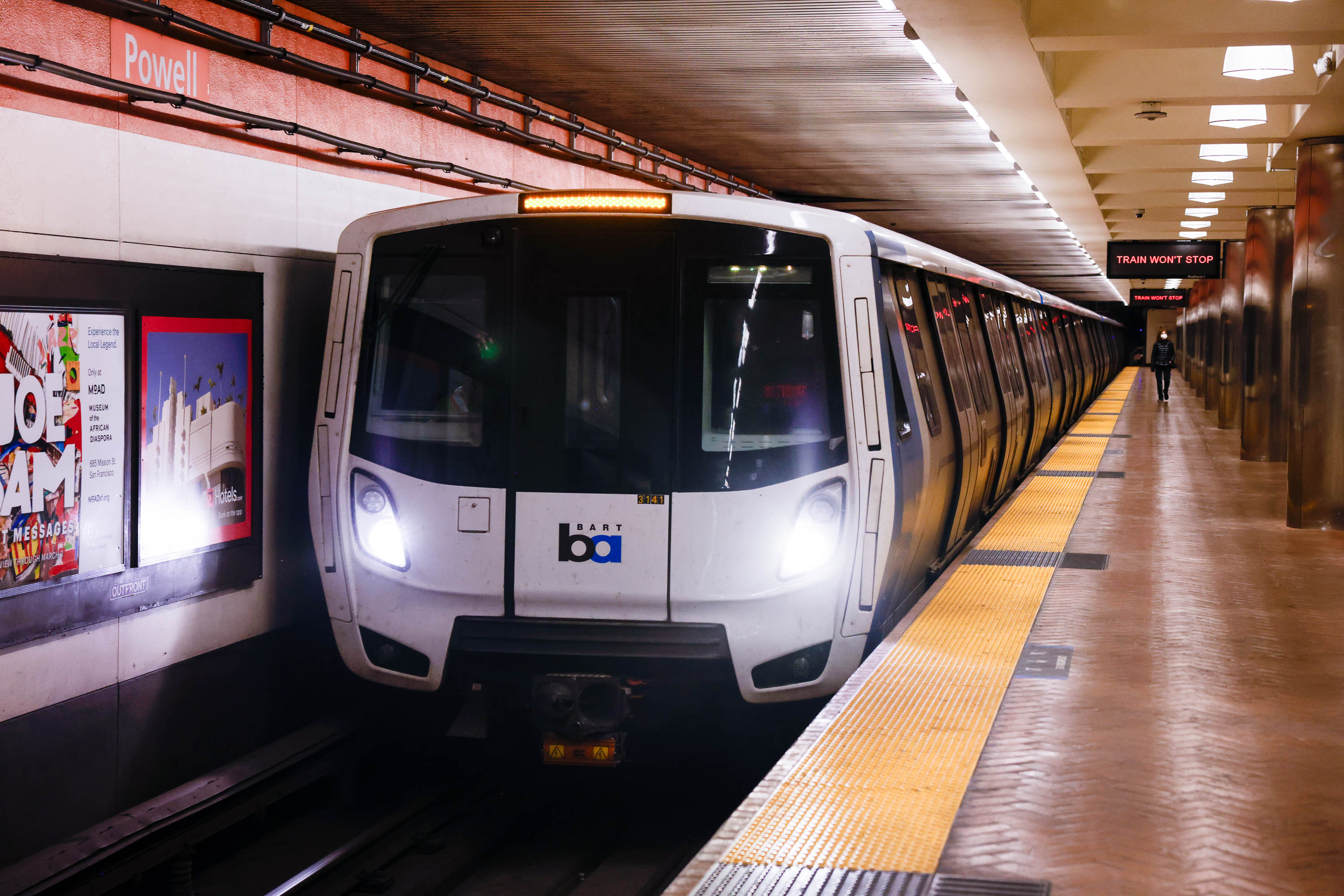 A BART train is at the Powell Street station, with its white front and headlights on. The platform has yellow tactile paving and posters on the walls.