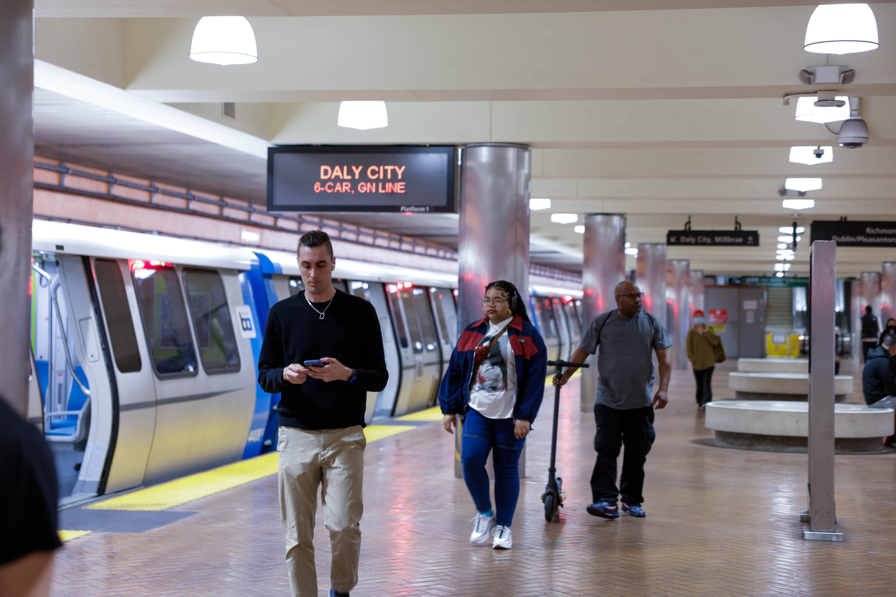 People are walking in a subway station with a train on the left. Above, a sign reads "Daly City, 8-Car, GN Line." The station is well-lit and busy.