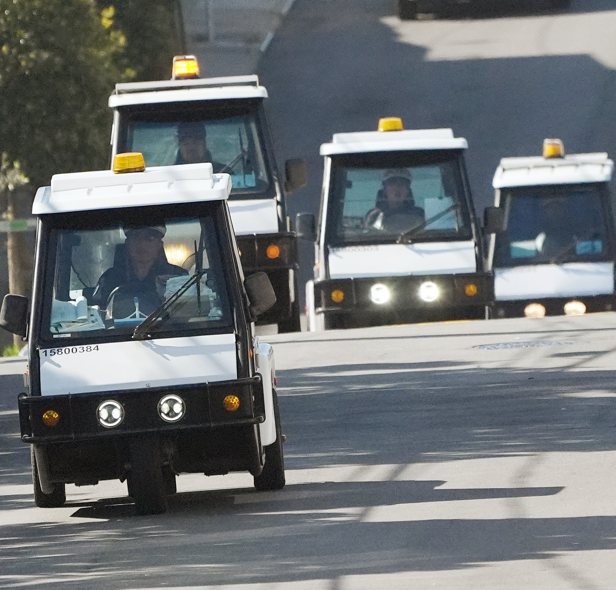 A convoy of small white utility vehicles, each with an amber light on top, drives down a road.
