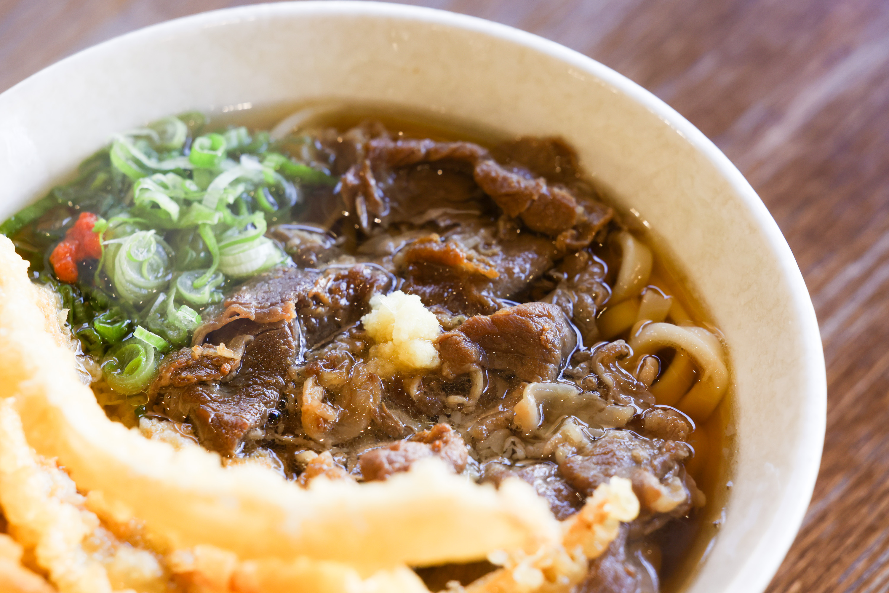 A bowl of beef udon with tempura, garnished with green onions and red spices.