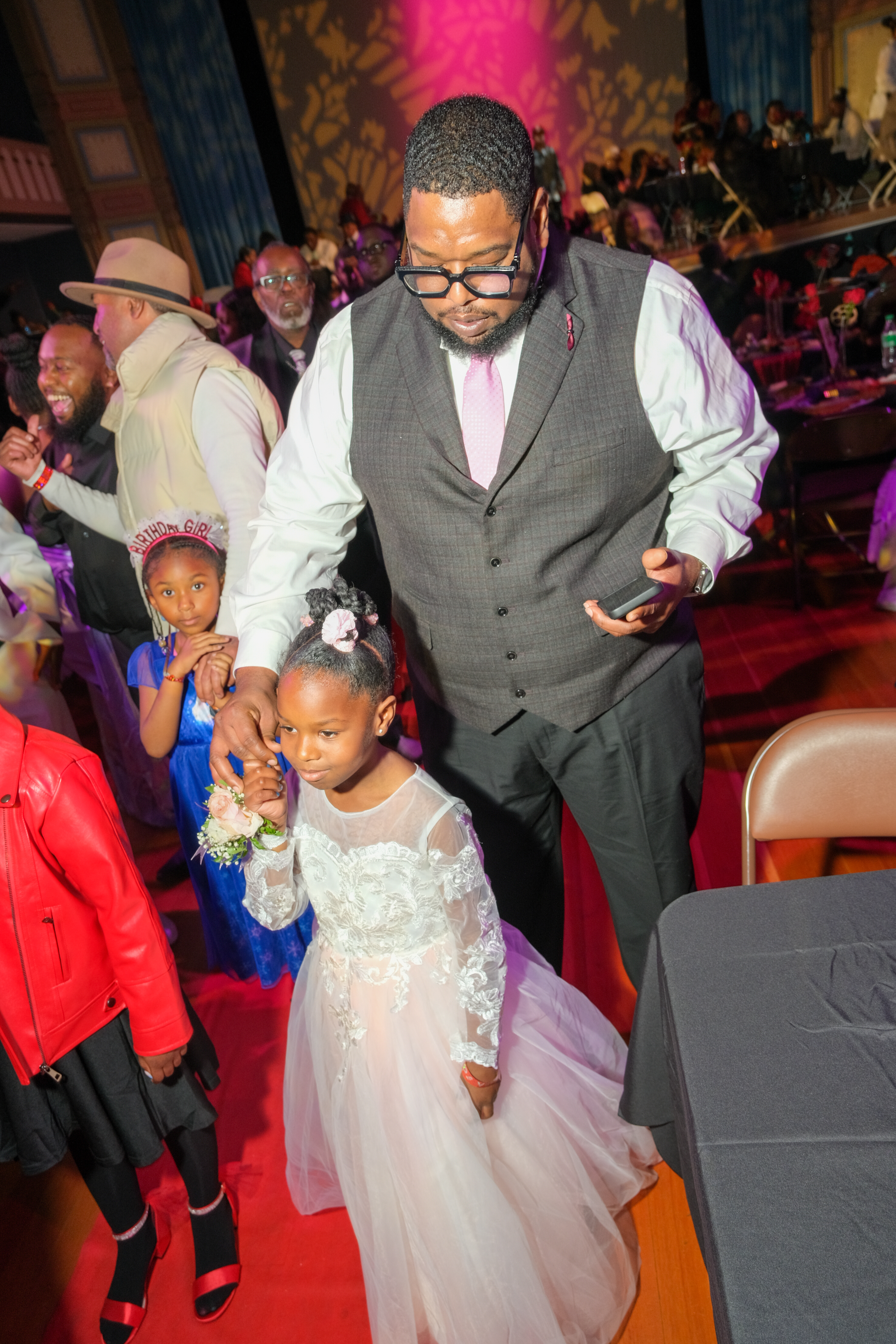 A man in a suit dances with a young girl in a white dress at a vibrant party setting with onlookers.
