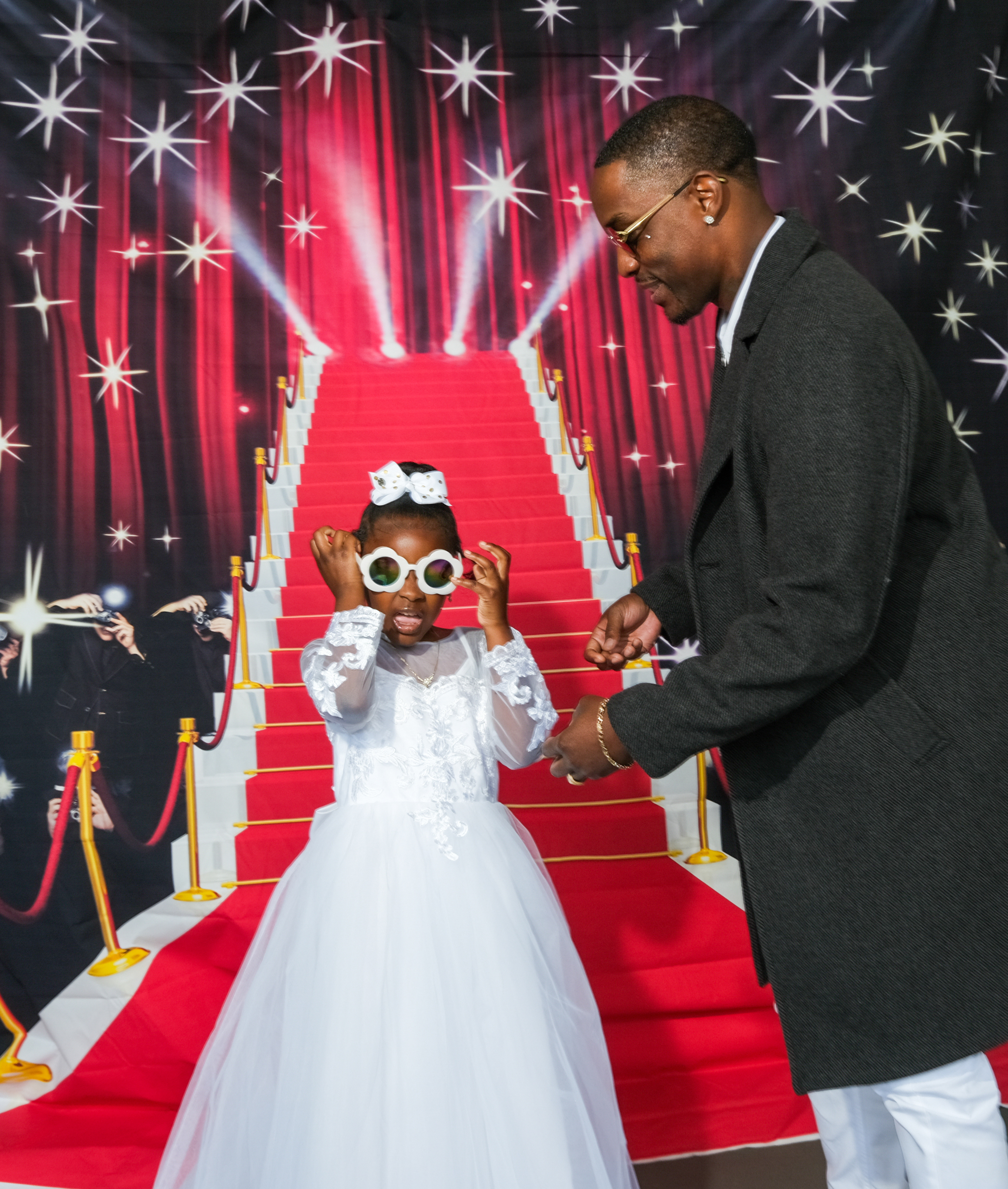 A young girl in a white dress adjusts big sunglasses, with a man beside her against a backdrop of a red carpet stairway and sparkling stars.
