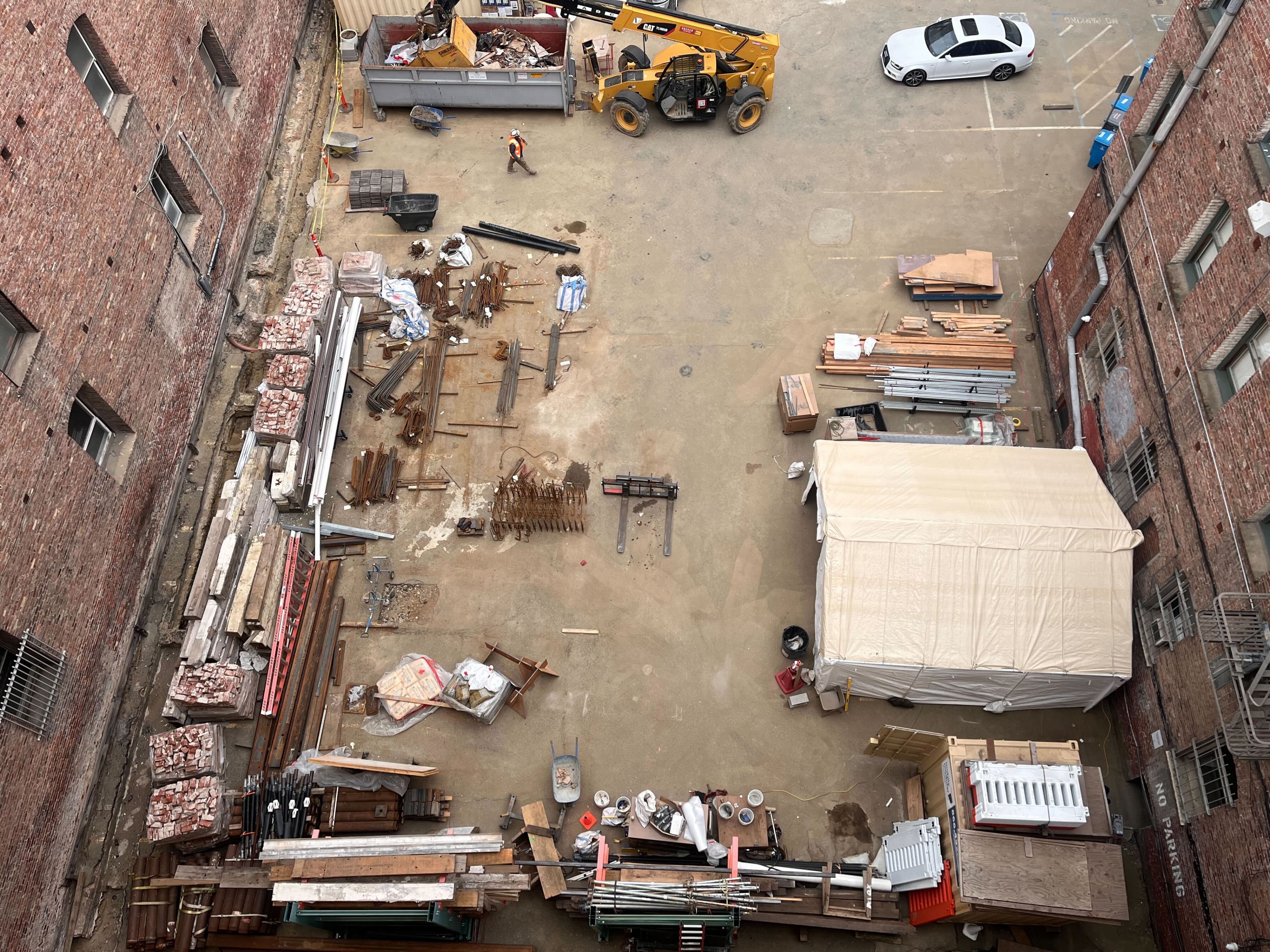 An aerial view of a construction site with materials, machinery, and a parked car, flanked by brick buildings.