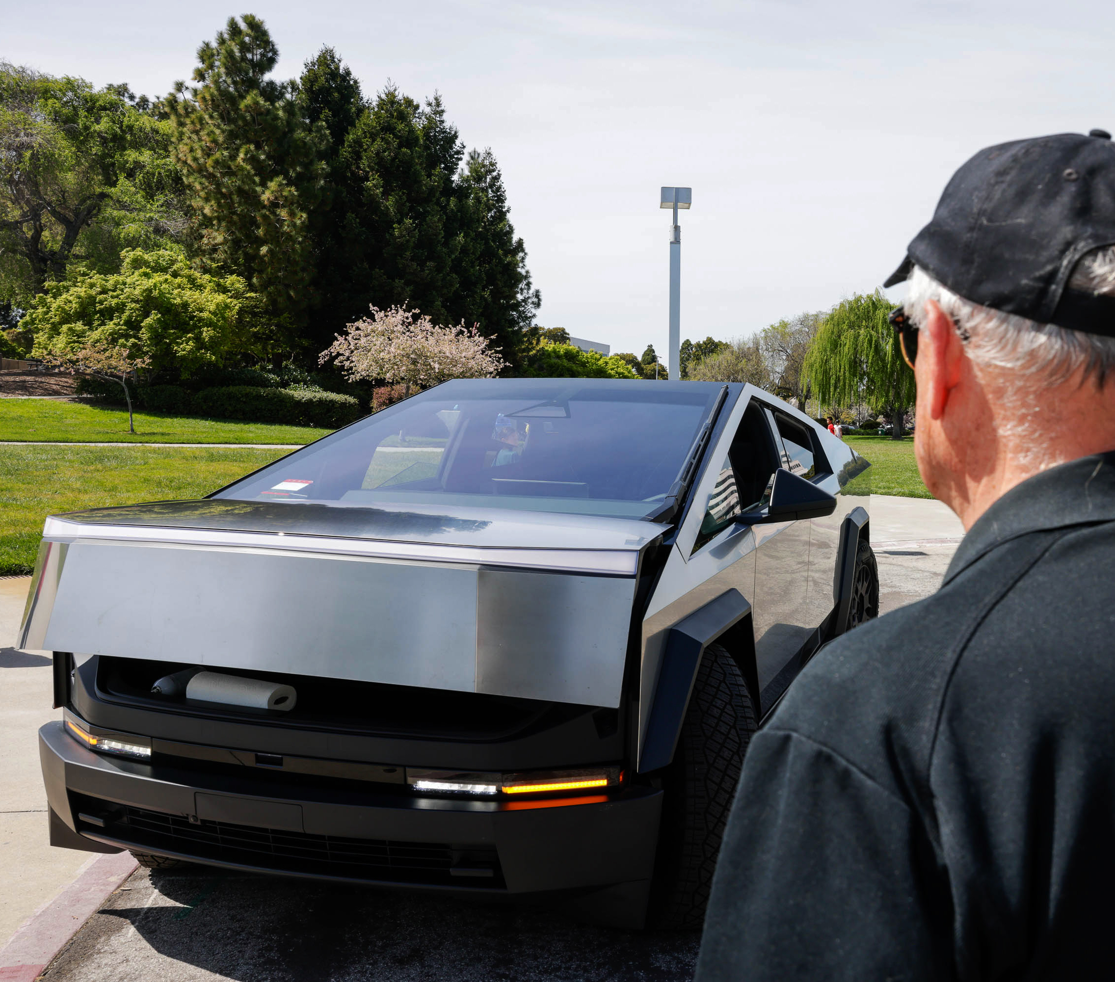 A man in a cap gazes at a silver, angular car with its gull-wing door open, parked outdoors.