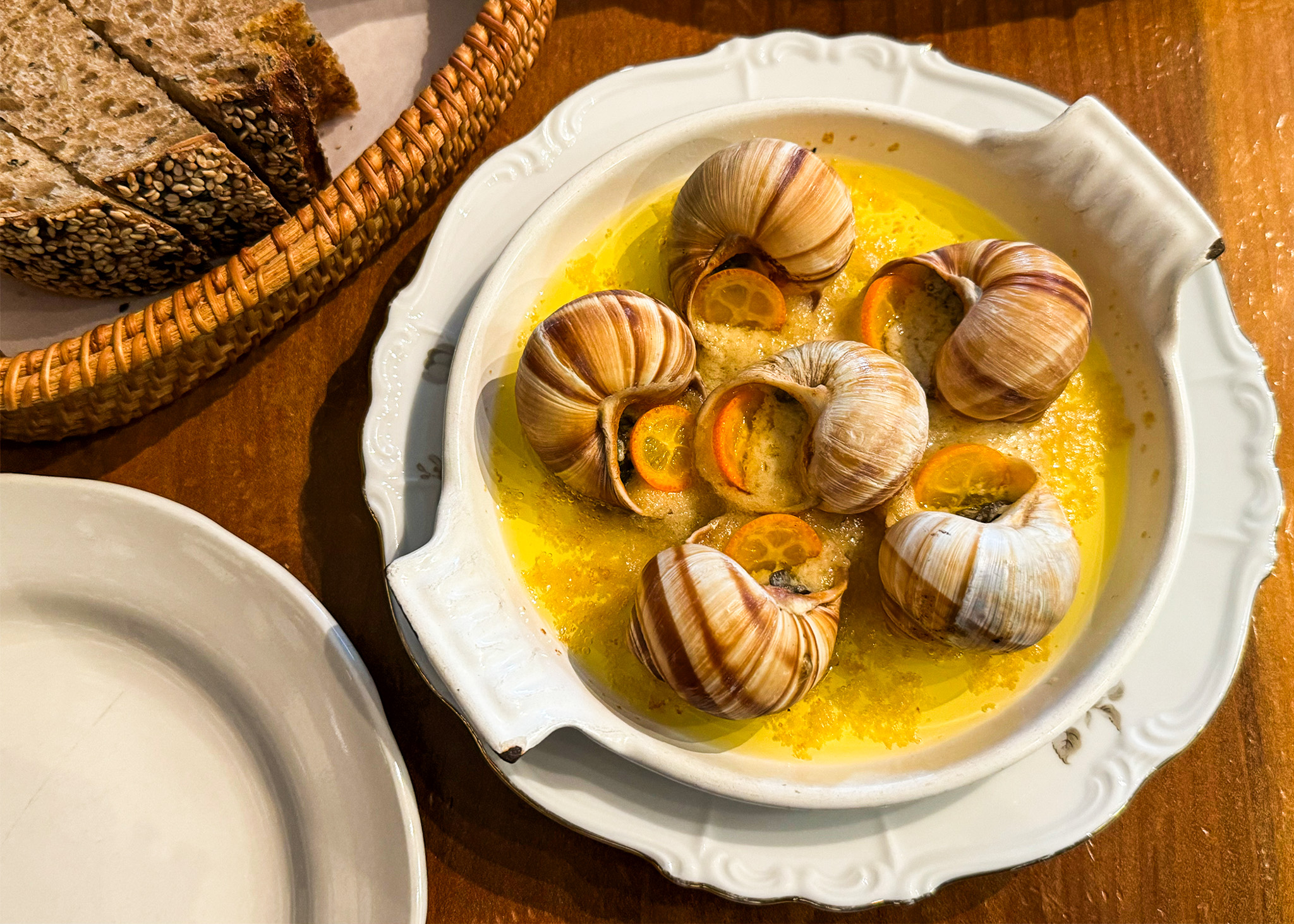A plate of escargot with garlic butter, slices of rustic bread, and an empty plate on a wooden table.