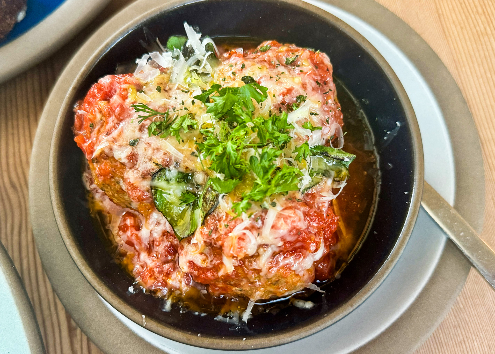 A baked dish topped with cheese and herbs, in tomato sauce, presented in a round bowl with a fork.