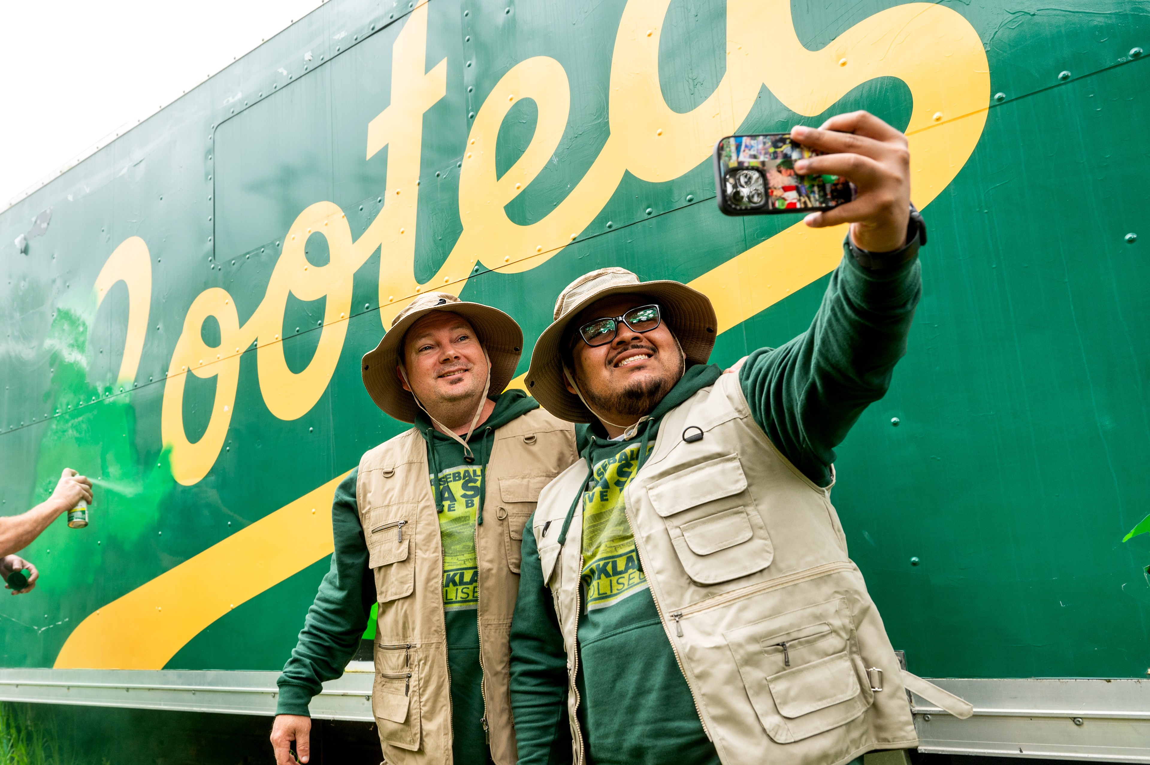 Two men in hats and vests take a selfie in front of a green truck with yellow text.