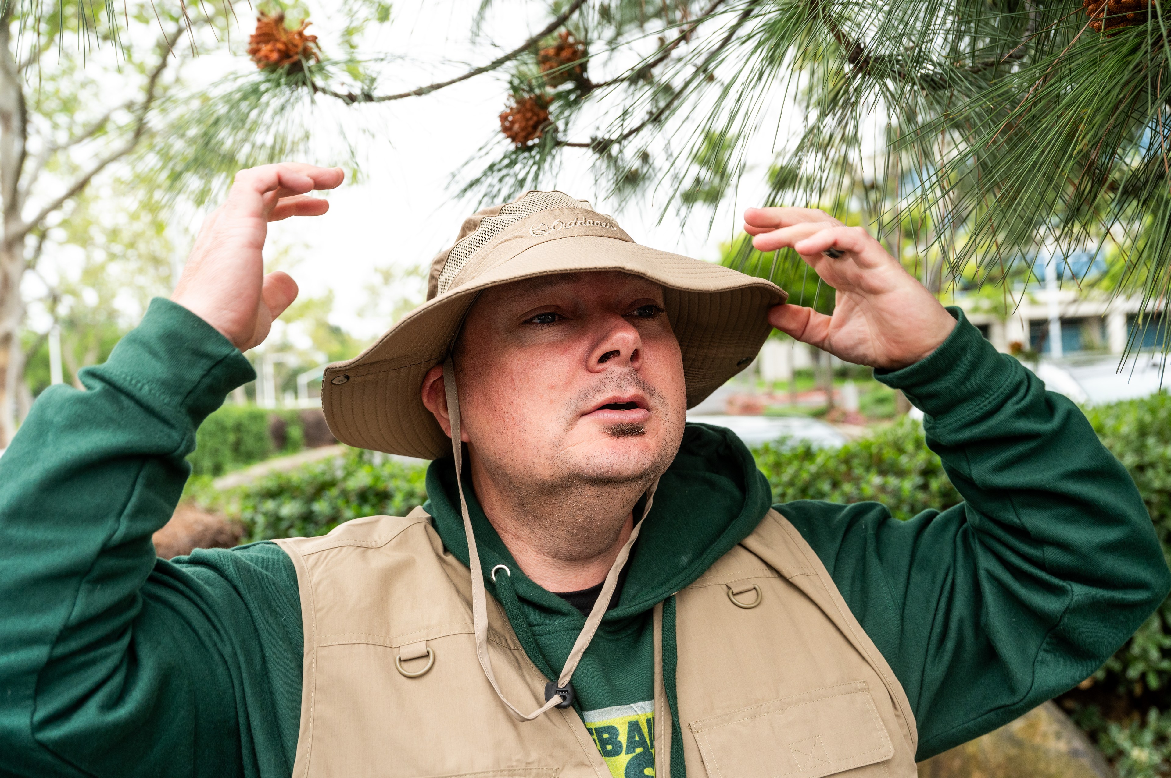 A man in a beige hat and green jacket looks up, touching the brim of his hat, with pine branches above.