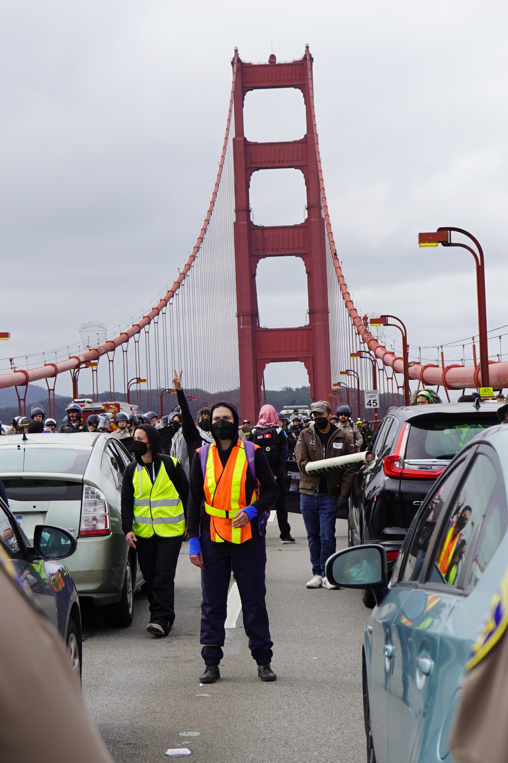 Police facing a line of stopped cars on the Golden Gate Bridge, with onlookers and emergency personnel.