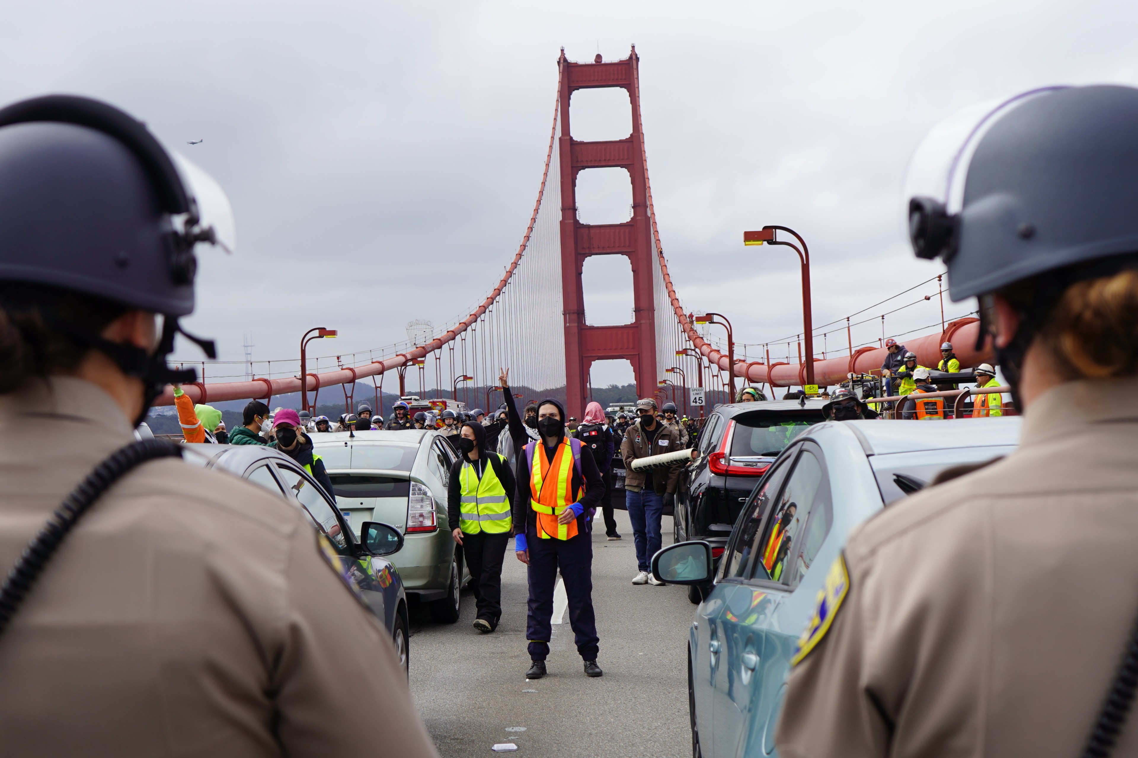 Police facing a line of stopped cars on the Golden Gate Bridge, with onlookers and emergency personnel.