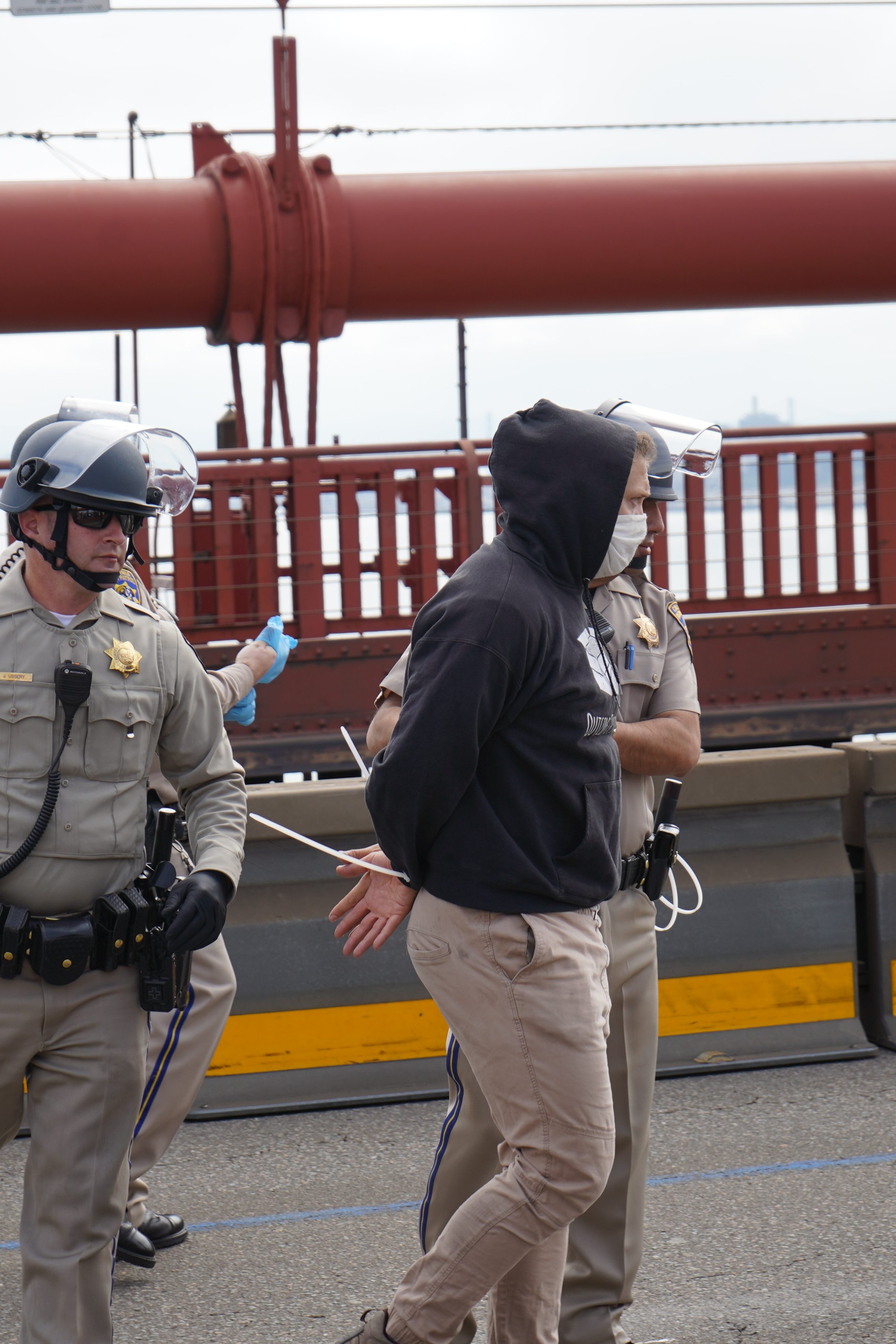 Two police officers in helmets handcuff a person in a hoodie on a bridge, with a thick red metal beam in the background.