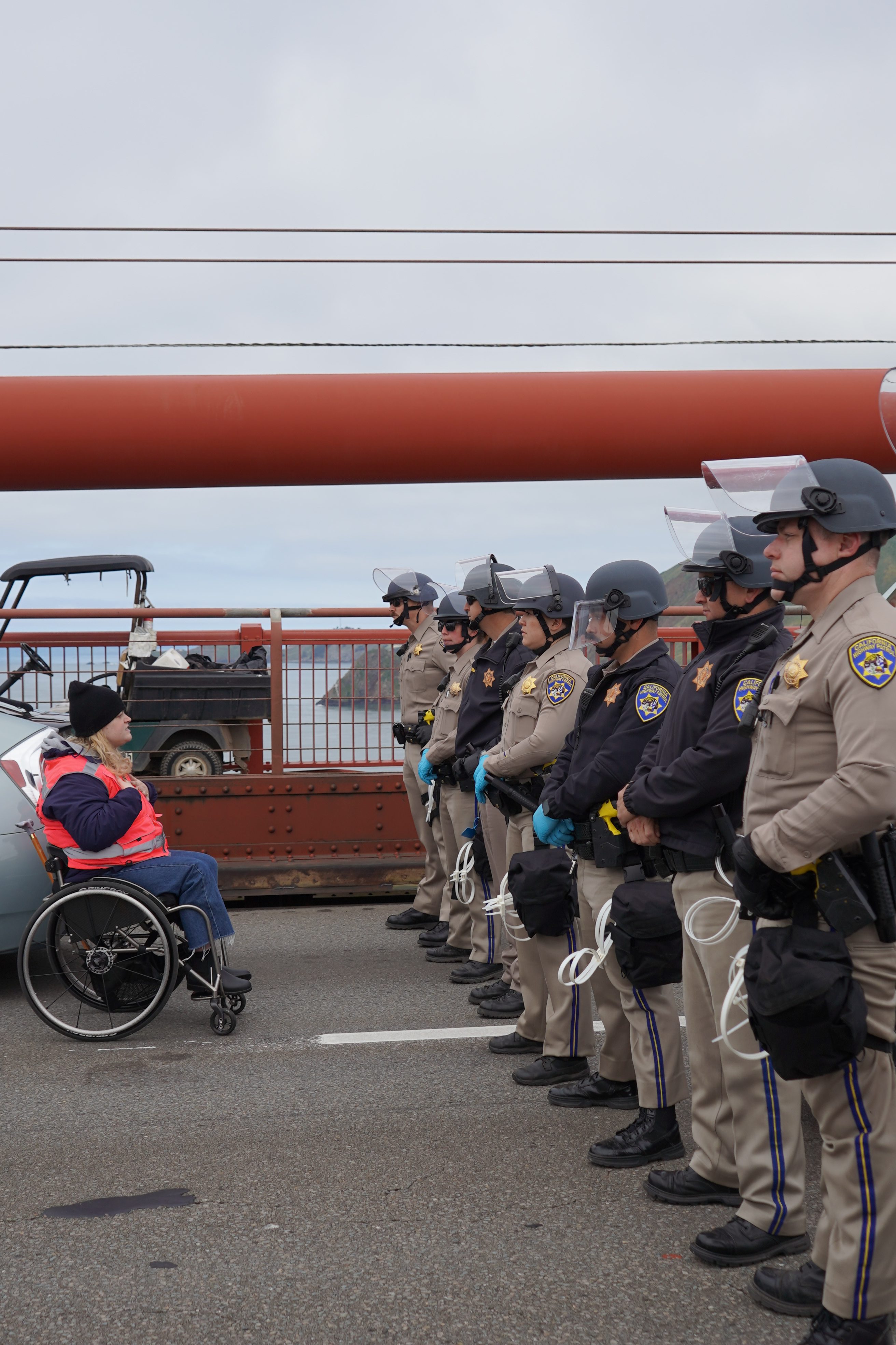 A person in a wheelchair wearing a bright orange vest faces a line of police officers in helmets and face shields on a bridge, with the background showing some water and distant land.